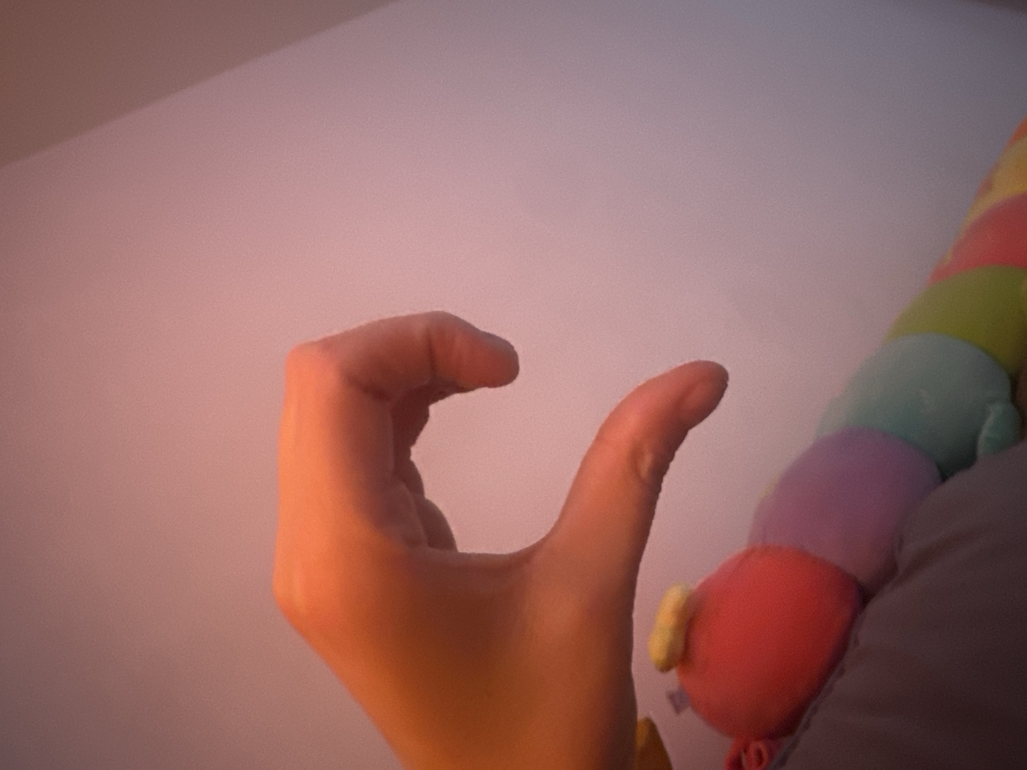 The image shows a close-up of a person's hand making a "C" shape. The hand is illuminated by warm light, casting shadows that highlight the contours of the fingers and palm. To the right of the hand, a colorful stuffed caterpillar toy is visible, with segments in red, pink, purple, green, and light blue. The background is a softly lit, textured wall, possibly a ceiling or a wall in a room. The lighting suggests it could be early morning or late afternoon, with a warm, ambient glow. The scene appears to be indoors, in a domestic setting. There is no visible text in the image. The overall mood is calm and relaxed, with the focus on the hand gesture and the soft textures of the objects.