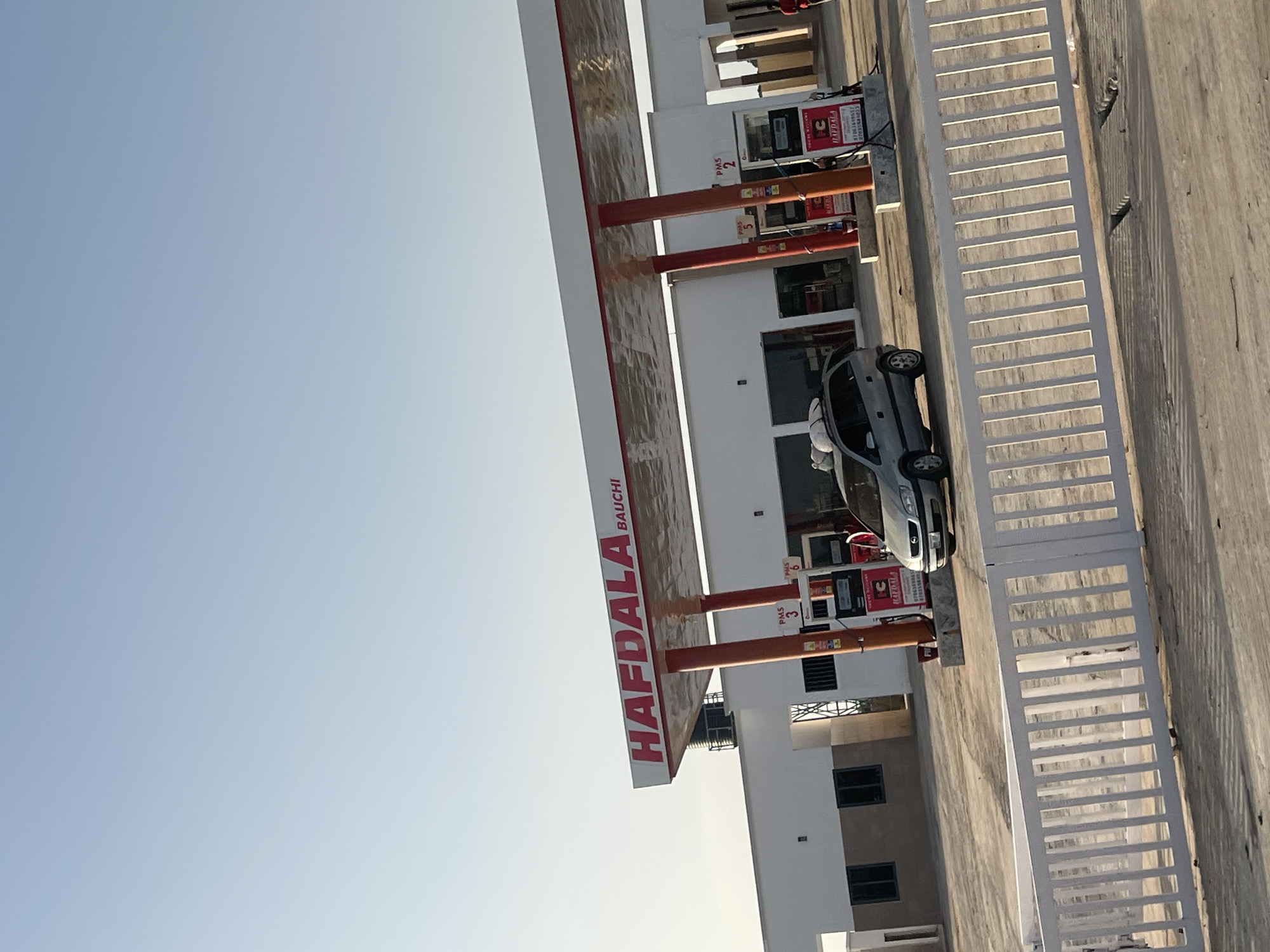 This image captures the Hafdala Bauchi gas station in Bauchi, Nigeria, under a clear, light blue sky. The gas station features a white building with a prominent red-trimmed canopy that displays "HAFDALA BAUCHI" in red lettering. Four fuel pumps are visible, supported by orange pillars, with labels including "PMS 3," "PMS 5," "PMS 6," and "PMS 2." One pump further displays "HAFDALA" branding along with phone numbers "07063584857" and "07031384033." A silver minivan with cargo secured to its roof rack is parked beneath the canopy, near the pumps. A single individual in a red shirt is partially visible on the far right of the frame, walking away from the pumps. In the foreground, a white, slatted metal gate or fence extends across the width of the image. The ground is a light-colored paved surface, exhibiting shadows cast by the fence, indicative of strong sunlight. A black water tank is visible on the roof of the building behind the canopy structure.