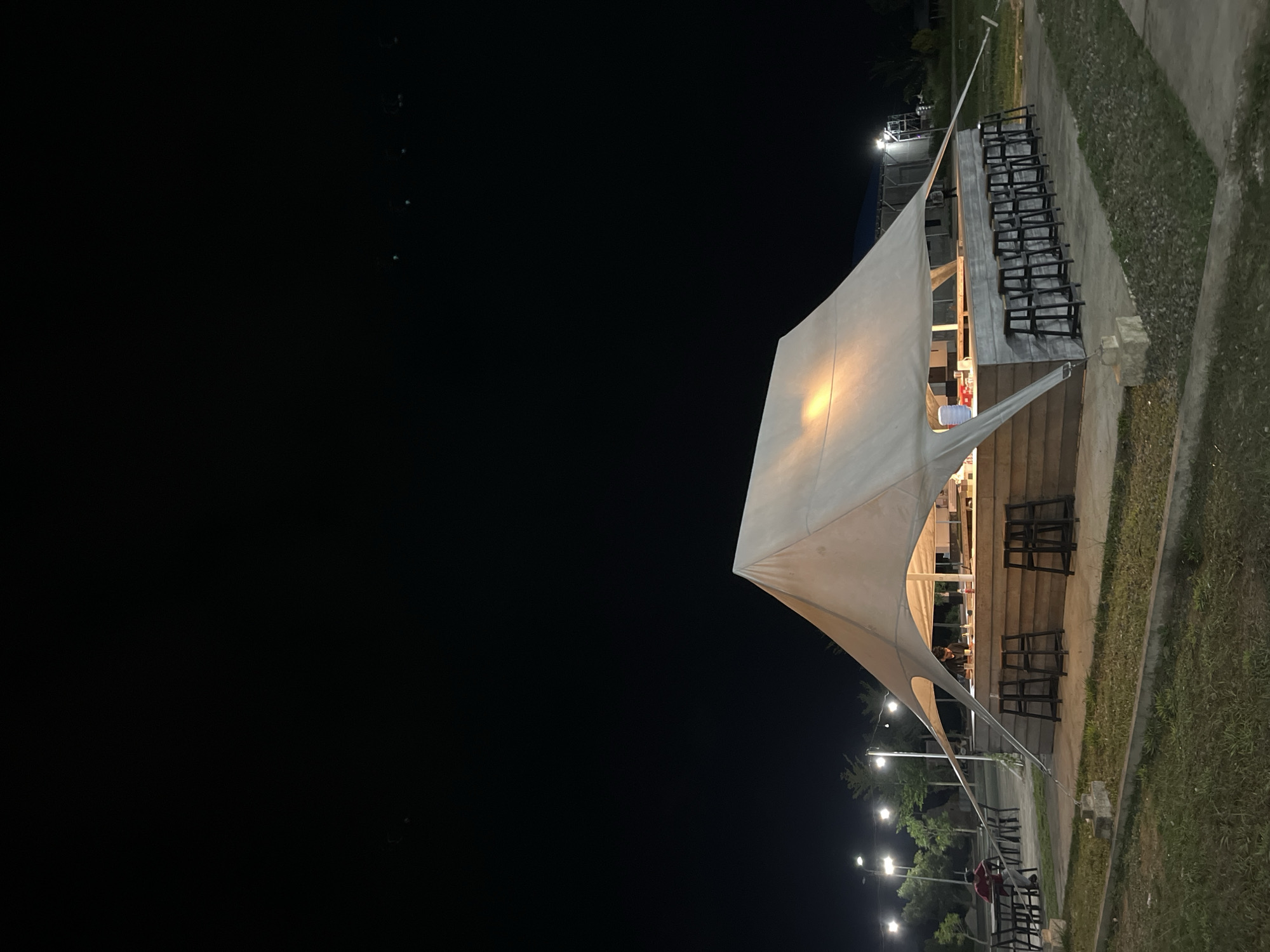 The image depicts a nighttime scene in Lhokseumawe, Indonesia, showcasing an outdoor structure, possibly a cafe or seating area, with a large white tent-like roof illuminated from within. Several rows of black metal chairs are arranged on concrete tiers beside the structure. The background consists of a dark sky, a row of evenly spaced streetlights along a path, and some trees. To the right, there is a grass-covered area and a concrete wall.