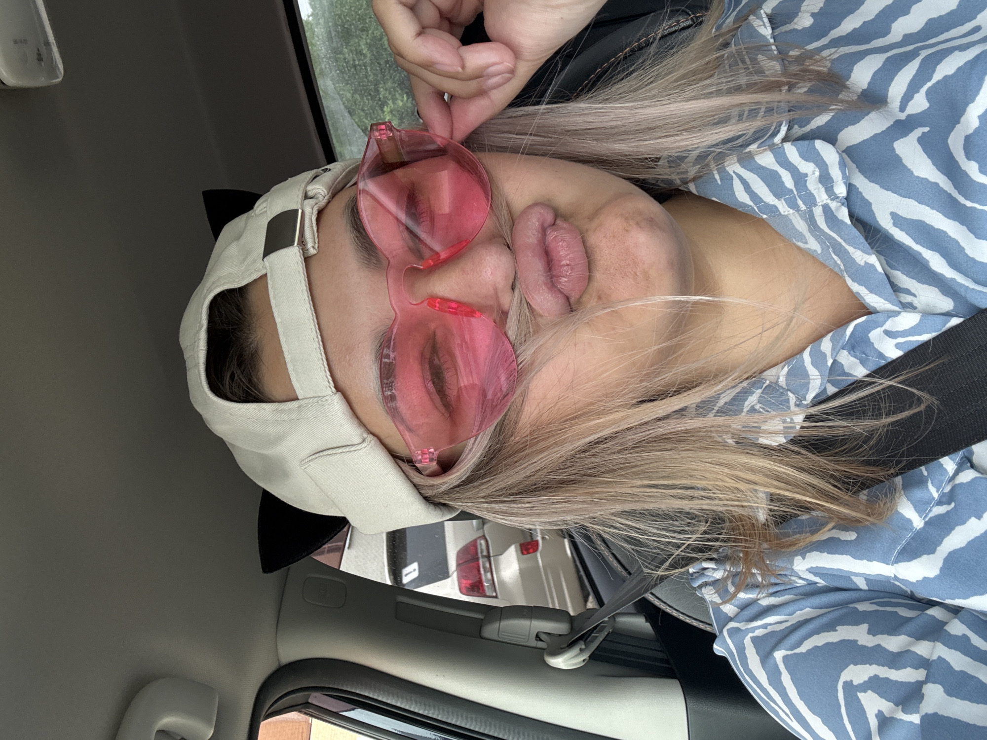 A young female with blonde hair is sitting in the driver's seat of a car, wearing a beige baseball cap with black cat ears attached. She has on pink heart-shaped sunglasses and is puckering her lips, holding a strand of her hair near her mouth. Her shirt has a blue and white zebra-like pattern. The scene appears to be in Birobidzhan, Russia, with a car visible through the rear window.