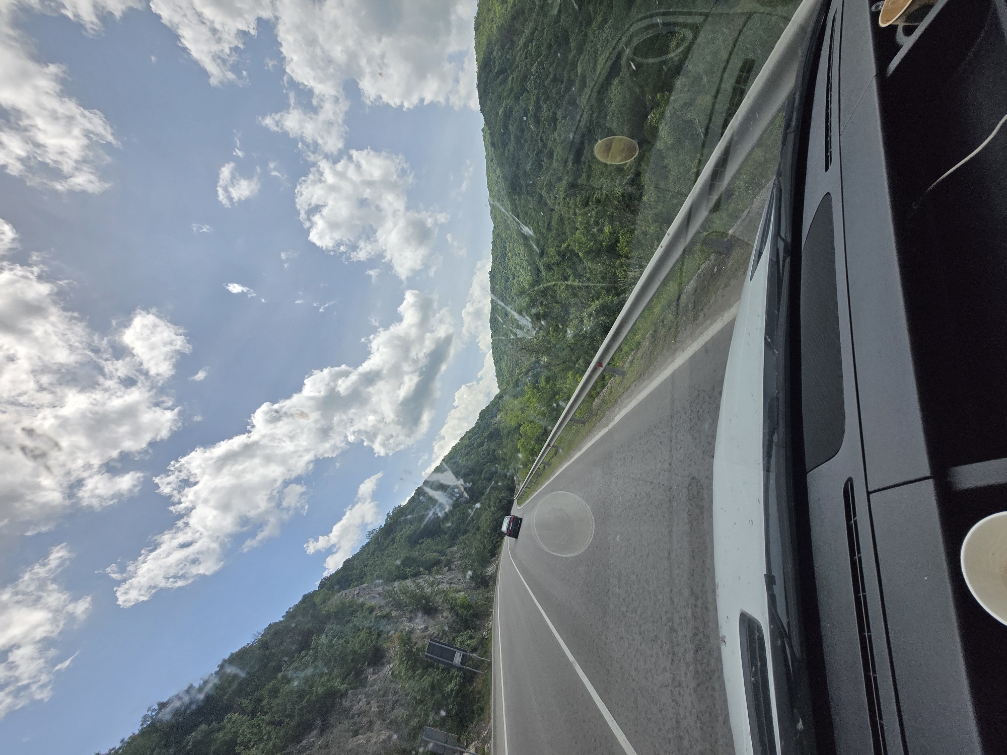 The image captures a scenic view from inside a vehicle traveling on a paved road through a mountainous region. The perspective is looking out the front passenger-side window, with the image rotated 90 degrees counter-clockwise, making the road appear vertical.
When correctly oriented, the road curves gently to the right in the mid-ground. A silver guardrail runs along the right edge of the road, bordering a steep, densely forested mountain slope. The background features a vast expanse of verdant, tree-covered mountains stretching across the horizon.
The left side of the image is dominated by a bright blue sky filled with numerous fluffy white clouds, indicating clear, sunny daytime weather, likely morning or afternoon.
In the immediate foreground, to the right, parts of the vehicle's interior are visible, including a dark dashboard or console. A white paper cup is partially seen in the bottom right corner. The vehicle's exterior, specifically a white section of the hood or fender, is visible through the windshield. Reflections and smudges are present on the glass. A small, dark-colored car can be seen in the distance on the road ahead, traveling in the same direction.
The scene depicts a peaceful journey through a lush, natural landscape. No text is visible in the image.