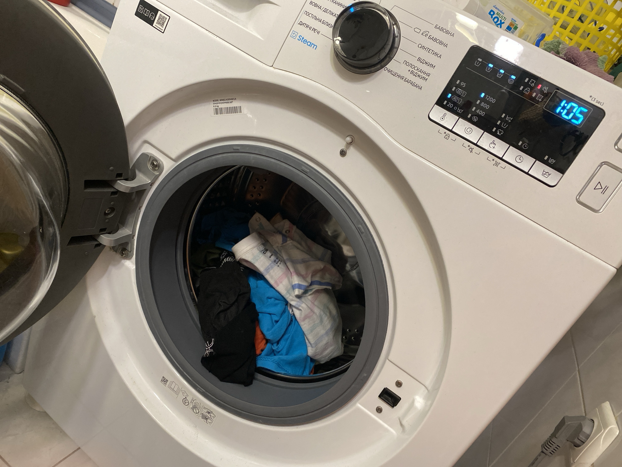 The image shows a white front-loading washing machine with its door open, revealing a load of laundry inside. The laundry appears to be mixed colors, including black, blue, orange, and striped items. The washing machine is situated in what appears to be a domestic setting, possibly a laundry room or bathroom, with tiled walls visible in the background. A power cord is plugged into a wall socket on the right side of the image. The washing machine's control panel, with a digital display showing "1:05" and various program options, is visible at the top right. This scene is located in the city of Kryvyi Rih, Ukraine.