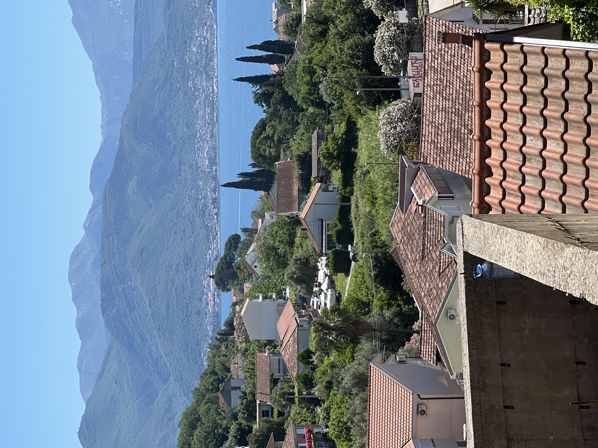 The image shows an elevated view of Baošići, Montenegro, towards the Bay of Kotor. The scene is dominated by the terracotta tiled roofs of houses nestled amidst lush green trees and foliage. In the distance, the calm blue waters of the Bay extend towards hazy, blue-grey mountains on the opposite shore. The foreground includes a concrete structure or wall, providing a sense of the elevated vantage point. There are no visible people, but the presence of houses suggests a residential area. The scene suggests a sunny, clear day.