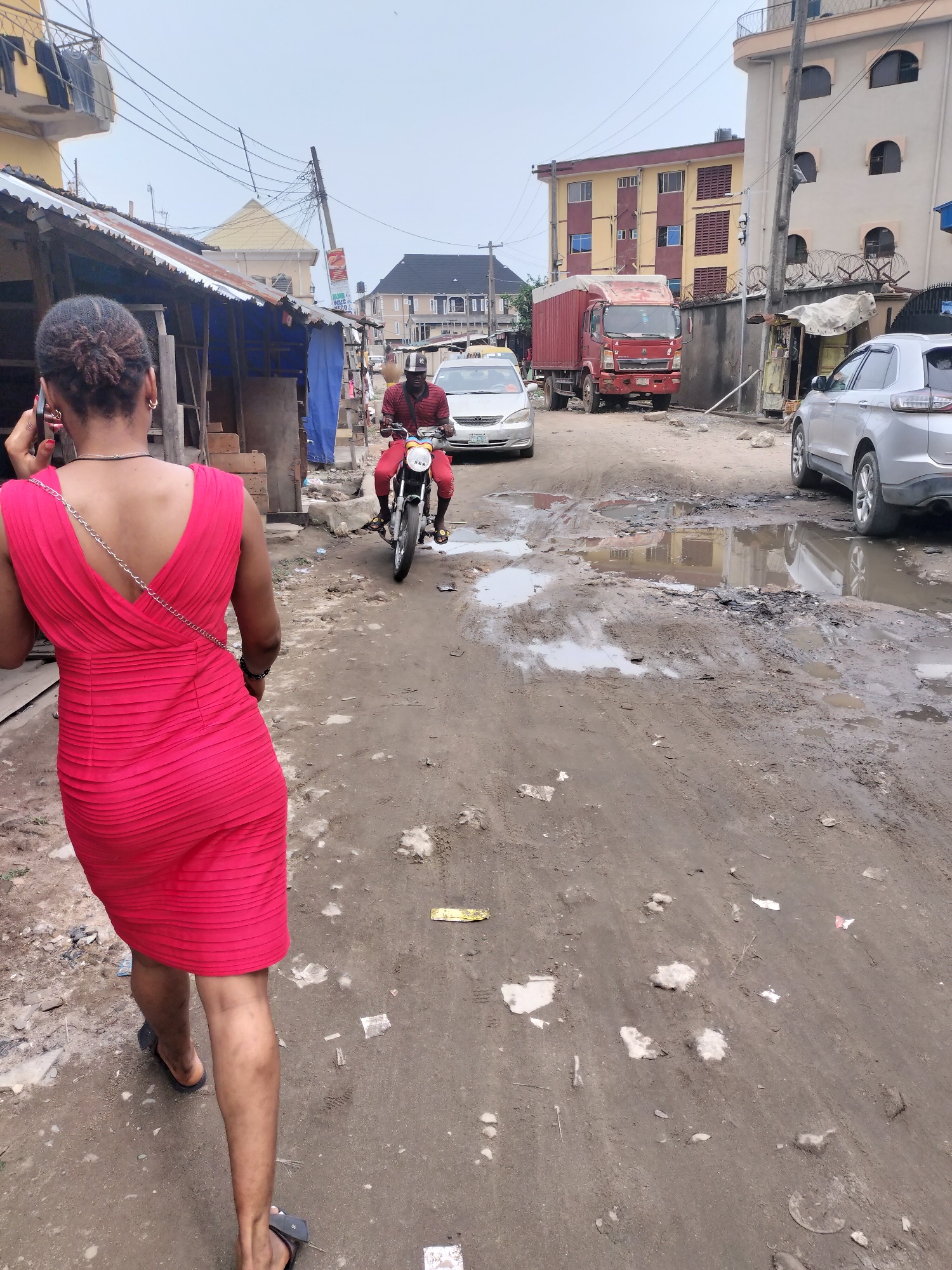 The image depicts a street scene in Alaiyabiagba, Nigeria. A woman wearing a red dress and sandals walks away from the camera while talking on a cell phone. A man rides a motorcycle towards the camera. Cars, a truck, and makeshift structures line the muddy, potholed street. Debris is scattered on the ground. The sky is overcast. Buildings are visible in the background.