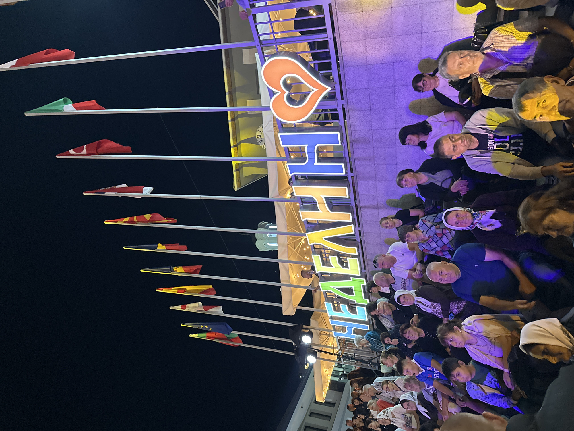 In Nedelino, Bulgaria, a crowd of people is seated outdoors at night, facing a brightly lit stage or performance area. A large, illuminated sign with letters and a heart shape is visible above the seating area. The sign appears to spell out "НЕДЕЛИНН" with the last letter being a stylized heart. Numerous flags of different countries are flying on poles behind the sign, suggesting an international event or celebration. The audience, composed of adults and children, is diverse in age and attire, with many looking towards the performance. The lighting is artificial, casting a purple and yellow glow on the scene, indicating it is evening. The background shows a building with awnings and a dome-like structure.