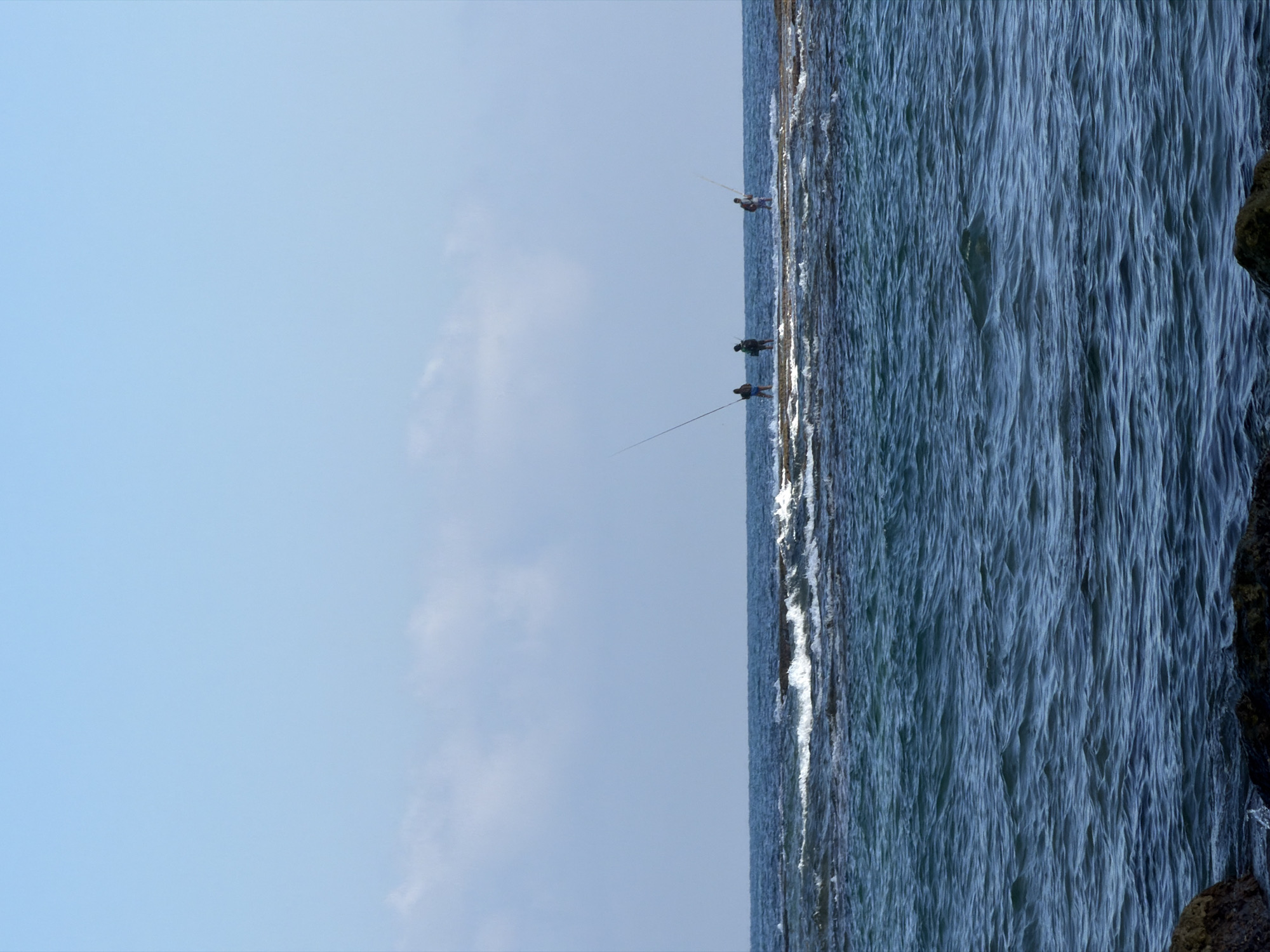 A bright, clear blue sky dominates the upper portion of the image. Below, a calm, blue sea stretches to the horizon, with a line of white-capped waves breaking over a submerged rocky reef. Three figures, appearing to be men, are visible standing on the reef, each holding a long fishing rod. They are spaced out along the reef, with the leftmost figure appearing to be the closest to the viewer. The foreground shows the dark, wet surfaces of rocks partially submerged in the water, suggesting the edge of a coastline or a rocky outcrop. The overall scene depicts a serene day of fishing in a coastal environment.