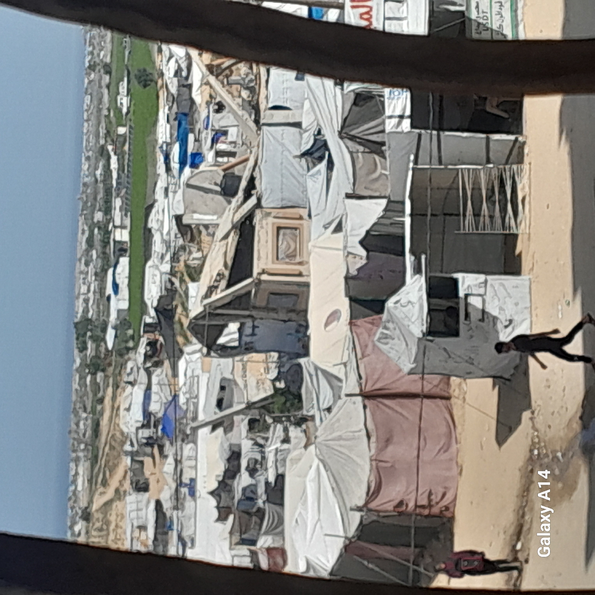Here is a detailed description of the image:
The image appears to depict a densely populated area with structures that resemble temporary or makeshift housing, possibly a refugee camp or informal settlement. The scene is viewed from an elevated perspective, partially framed by a dark foreground object.
On the left side of the image, there is a strip of land, possibly a field or vegetation along the edge of the settlement. The majority of the view is filled with a dense collection of shelters. These structures are varied, with some appearing to be tents or fabric-covered dwellings. The materials used for construction seem basic and readily available.
Two figures are visible in the lower right portion of the frame. One person is seen running, while another is lying on the ground.
Overall, the image conveys a sense of overcrowded conditions. The details suggest a challenging living environment characterized by limited resources and densely packed housing. The location is unknown.