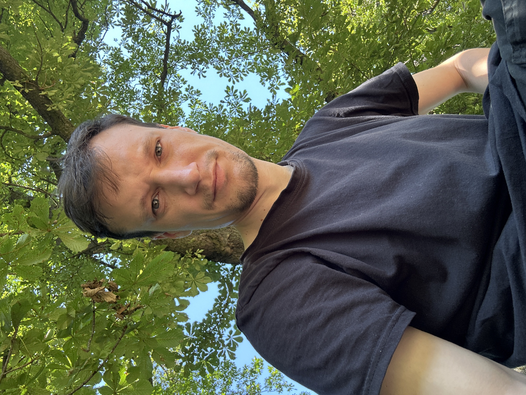 A close-up, low-angle selfie shows a man with fair skin, light brown hair, and a short beard. He is wearing a black t-shirt and appears to be outdoors, with lush green foliage and a blue sky visible in the background. The leaves suggest a temperate climate, possibly in a park or wooded area. The man's gaze is directed towards the camera, and his expression is neutral. There are no other people or objects clearly visible in the frame besides the man and the natural environment. The image does not provide any specific geographical markers or clues to indicate the location in Makiv, Ukraine.