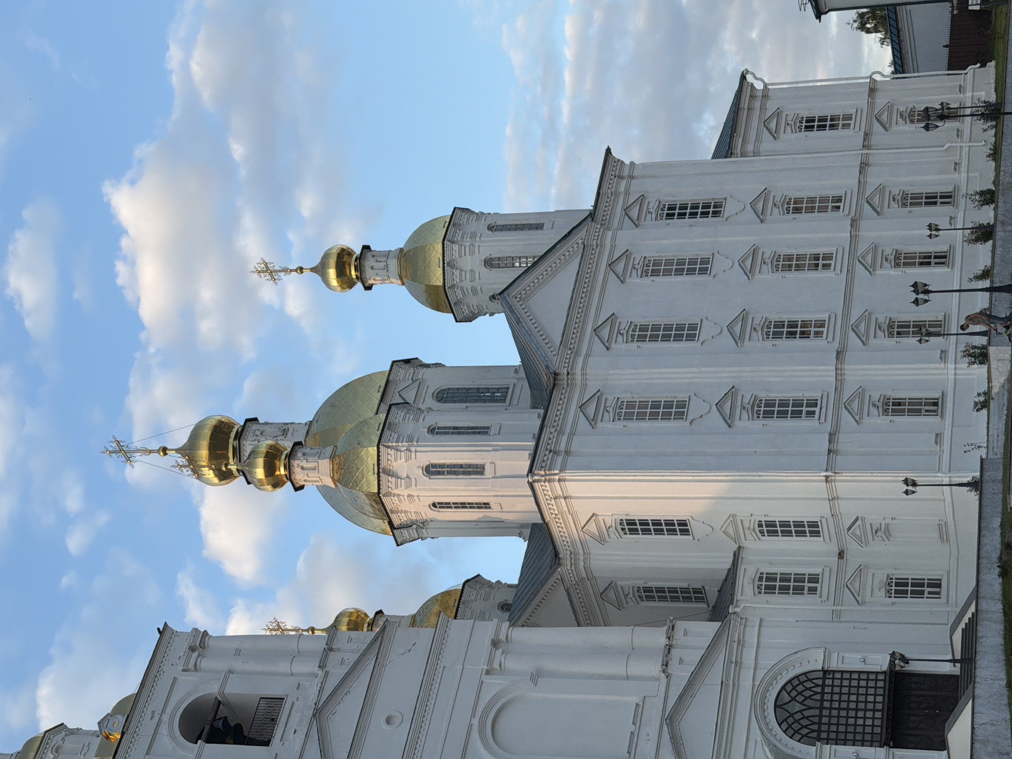 The image shows a view of the Cathedral of the Resurrection, located in the city of Arzamas, Russia. The white, multi-domed cathedral features golden domes topped with crosses. The sky above is blue with scattered white clouds, indicating daytime. The cathedral is viewed from a slightly elevated position, with some of the grounds and lampposts in the foreground. One lamppost is visible on the left, and several are visible on the right, closer to the building. A person is seen in the lower right quadrant of the image, tending to some plants near the lampposts. The architecture of the cathedral is ornate, with rows of windows and decorative elements on its facade. The overall scene depicts a religious landmark under a partly cloudy sky.