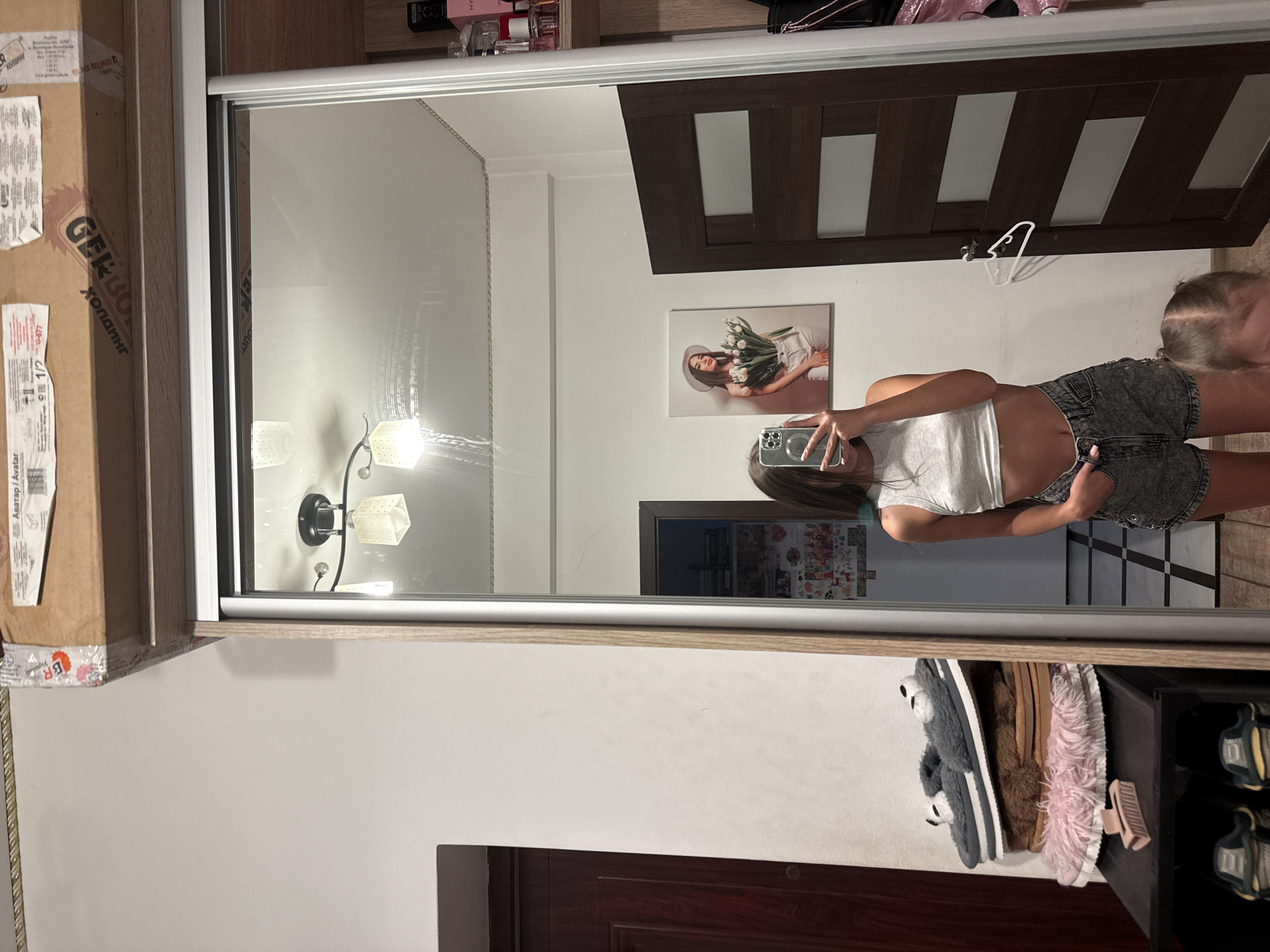 In this image, a young woman is taking a selfie in a bedroom. She is wearing a grey crop top and denim shorts. She is standing in front of a mirrored closet, and a young child is partially visible in the lower right corner of the frame. On a shelf to the left, there are several pairs of slippers and a shoe rack below. A framed picture of a woman holding tulips hangs on the wall to the left of the closet. The room appears to be tidy and well-lit, with a ceiling light fixture visible in the mirror. The presence of a wardrobe, shelf unit, and personal belongings suggests this is a residential setting. The background shows a door and more of the mirrored closet. The image is captured from a medium-full shot, focusing on the woman and her immediate surroundings.