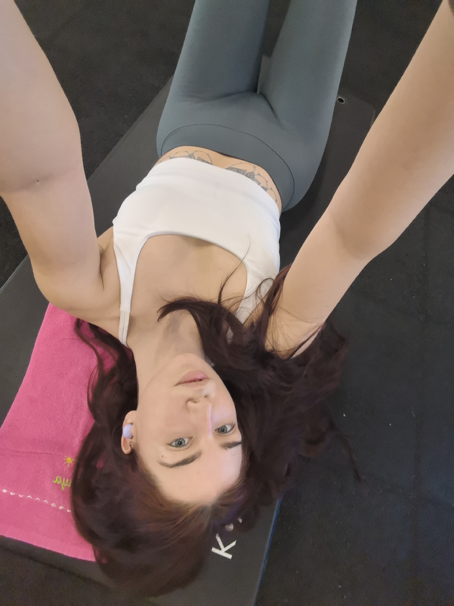The image shows a person lying on a yoga mat with their head tilted to the side, looking directly at the camera. They have long, dark brown hair that is spread out around their head. The person is wearing a white sports bra and dark grey leggings. A pink towel is positioned under their head. A single purple wireless earbud is visible in their right ear. The setting appears to be a gym or fitness studio, indicated by the dark, textured mat and the black floor. The lighting is bright, suggesting daytime or well-lit indoor conditions. The person's expression is neutral. Visible text on the yoga mat includes the letters "K" and "i", and on the pink towel, there is a yellow sun graphic and faint yellow text that is difficult to read but appears to be in Cyrillic. The overall impression is of someone resting or preparing for a workout.