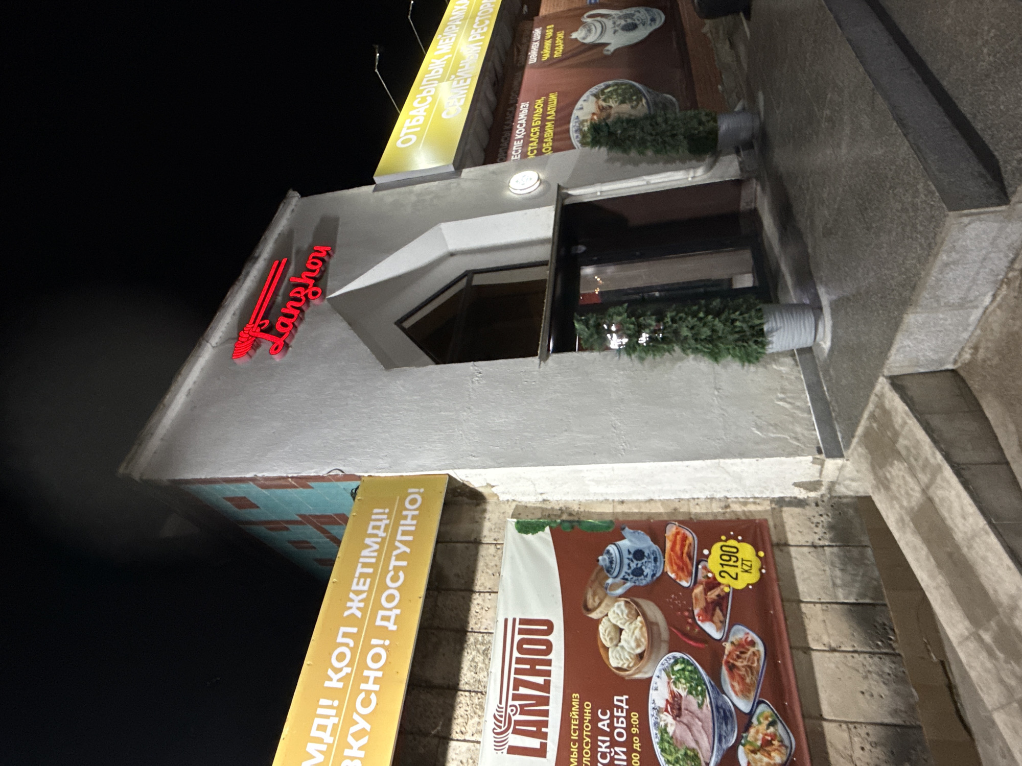 The image shows the exterior of the "Lanzhou" restaurant in Kokshetau, Kazakhstan, at night. The restaurant's name is displayed in red neon letters on the building. Yellow signs advertise the restaurant with phrases in both Kazakh and Russian. One sign translates to "Delicious! Affordable!" while another offers "teapot tea" as a gift. A large poster features various dishes with a price of 2190 KZT. Plants in gray planters are situated near the entrance. The architecture is a light-colored, modern design. The exterior includes textured stone or concrete.
