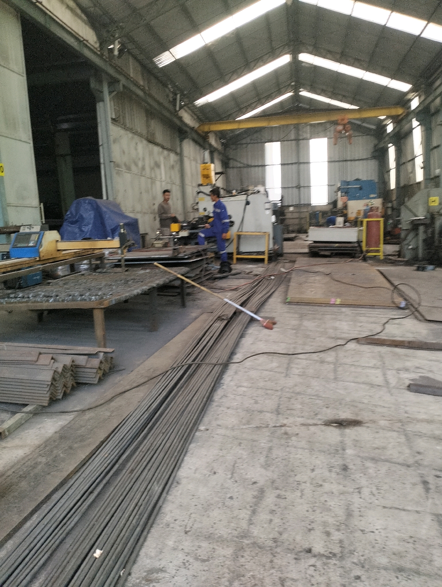 An interior view of a metal fabrication workshop in Gunung Sindur, Indonesia, shows a man in a grey shirt standing near a machine, and another man in a blue uniform operating a welding machine. The workshop floor is concrete, scattered with various metal objects, including stacks of rebar and flat metal sheets. In the background, industrial machinery and equipment are visible, with a large overhead crane present in the ceiling. The space is illuminated by natural light coming from the translucent panels on the roof and some artificial lighting.