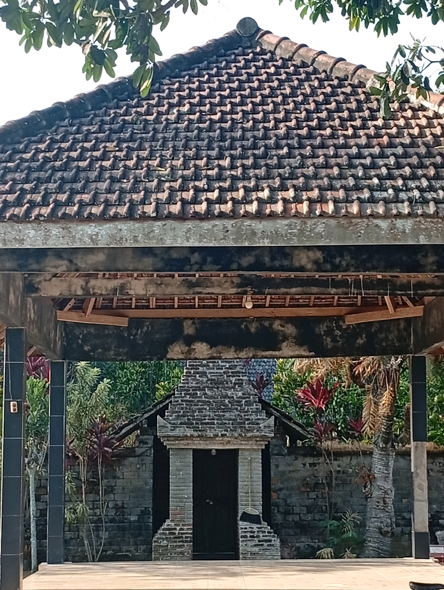The image depicts an outdoor scene in the village of Wajak Kidul, Indonesia. In the foreground, there is a tiled roof structure with visible wooden supports underneath. Below this, a path or courtyard is visible, leading to a brick building. This building features an entrance with a dark door, framed by brickwork. The area around the building is adorned with various plants, including trees and bushes with red and green foliage. The overall impression is of a traditional Indonesian architectural style, set within a natural environment.