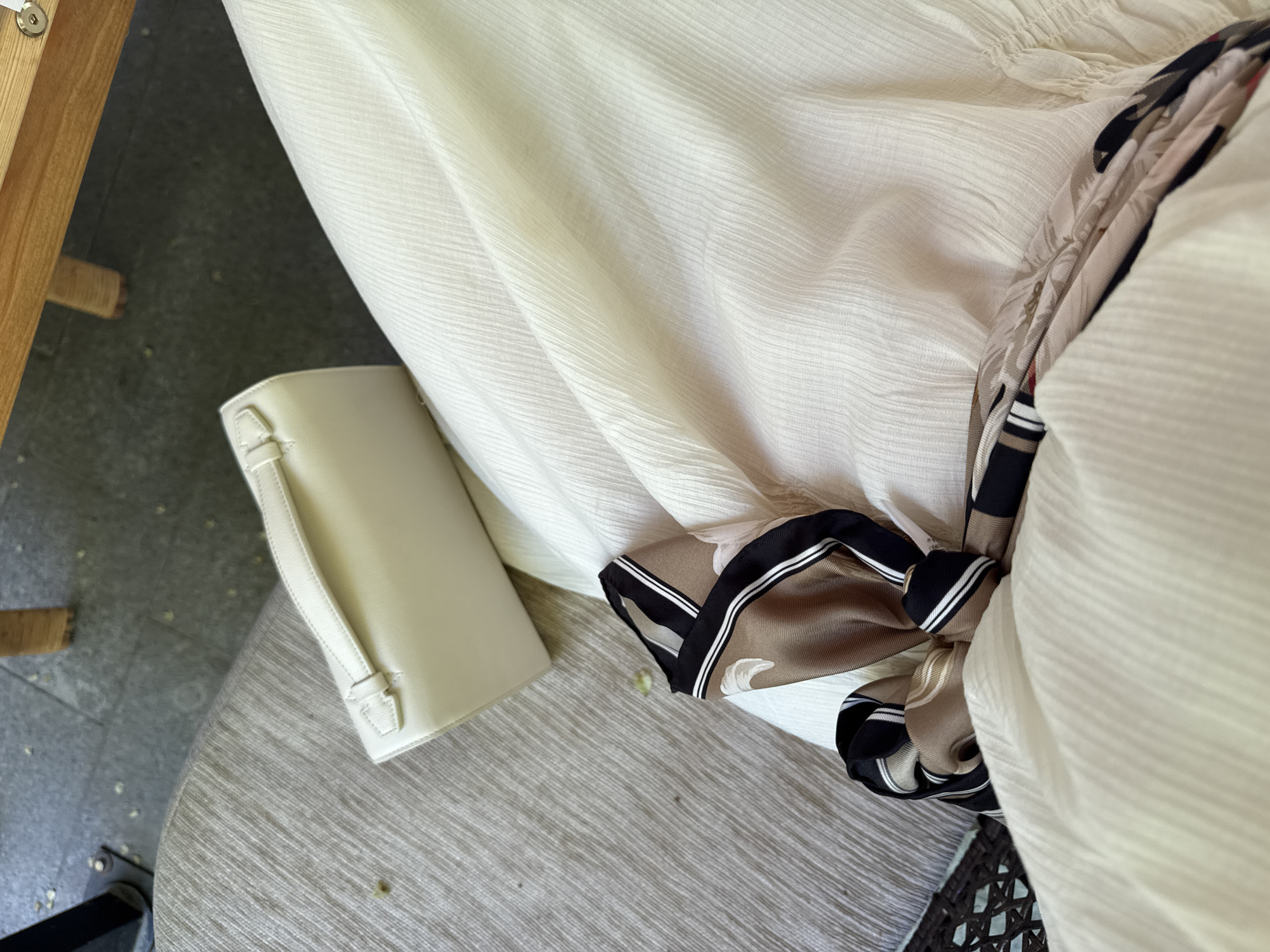 The image shows a close-up of a light-colored dress or piece of fabric draped over a surface. Next to it, a small, light-colored handbag is visible. A patterned scarf with brown, black, and white stripes is partially visible near the edge of the fabric. The scene is indoors.