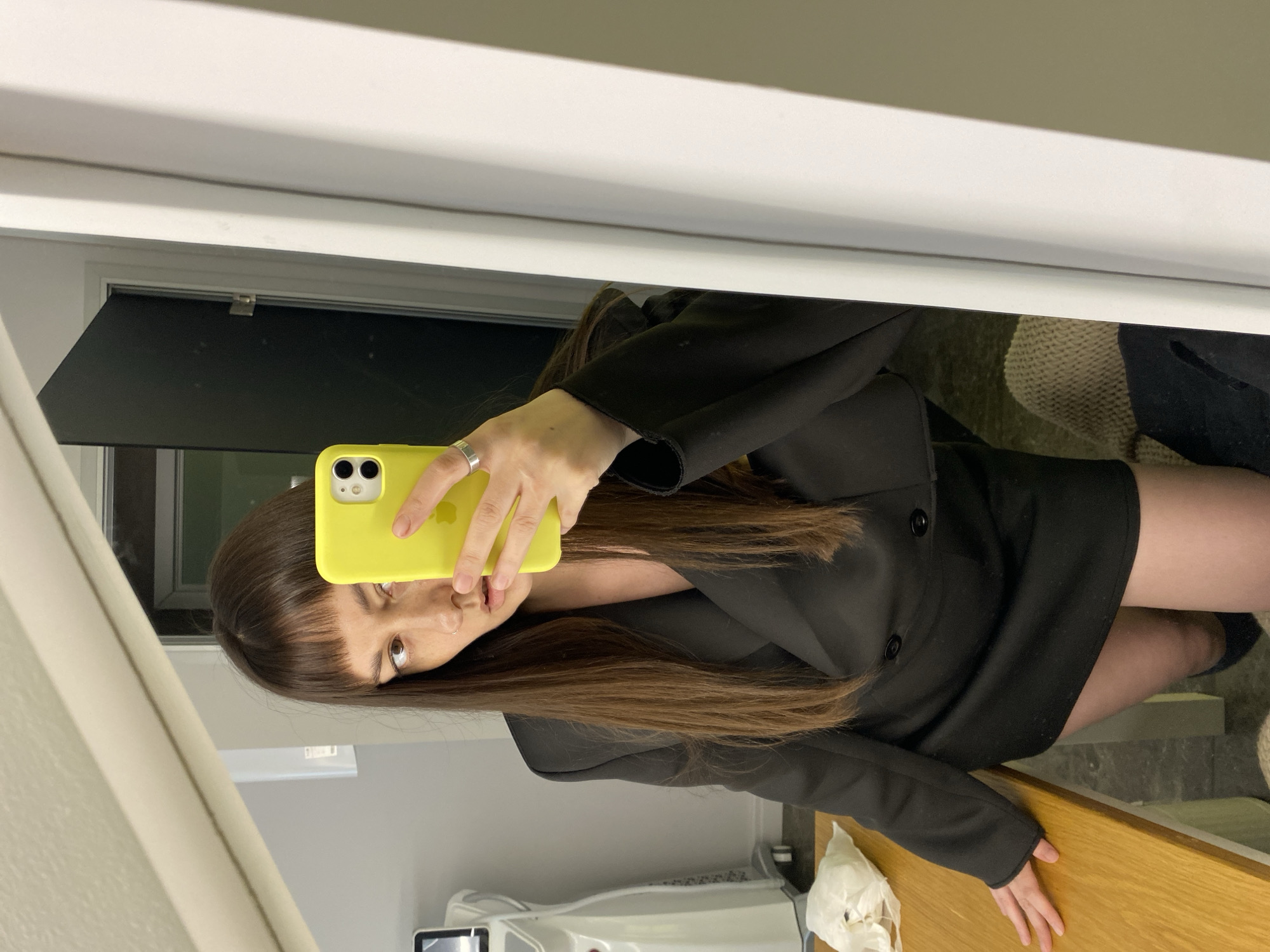 A woman is taking a mirror selfie with a yellow iPhone. She is wearing a black blazer dress. Her brown hair has bangs. The reflection shows a bathroom setting with gray walls and white trim. A window with a black shade is visible. The scene also includes a wooden countertop with crumpled tissues. The location cannot be determined to be specifically in Votkinsk, Russia based on the visual information provided.