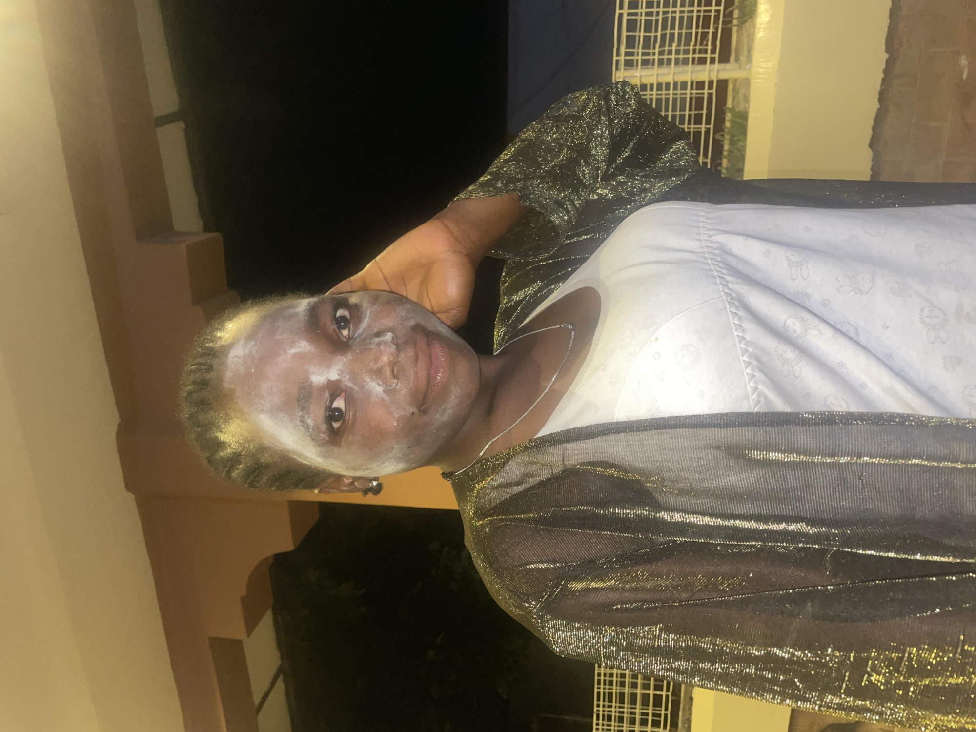 A woman with a white facial mask is posing in front of a building at night. She is wearing a white top and a gold mesh jacket. The location is likely in Jos, Nigeria.