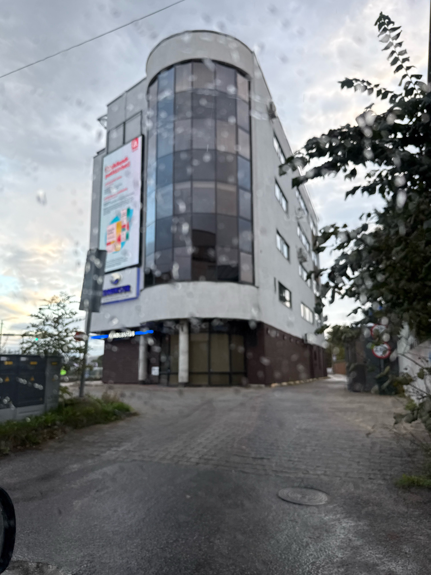A low-angle, slightly blurry exterior shot shows a modern, multi-story building with a curved glass facade on the upper floors and a lighter colored exterior on the lower floors. A large, vertical advertisement with colorful graphics is mounted on the curved section of the building. Below the advertisement, a blue illuminated sign is visible on the ground floor. A small tree stands to the left of the building, and a dark-colored utility box is present in the foreground. The sky is overcast with clouds. The ground in front of the building is paved with cobblestones. To the right of the building, a tree with dark green leaves partially obscures the view. A red circular sign with black markings is visible near the entrance of the building. A sewer manhole cover is present in the paved area in the foreground. The overall impression is an urban environment, possibly in Tallinn, Estonia, with a focus on the architectural details of the building.