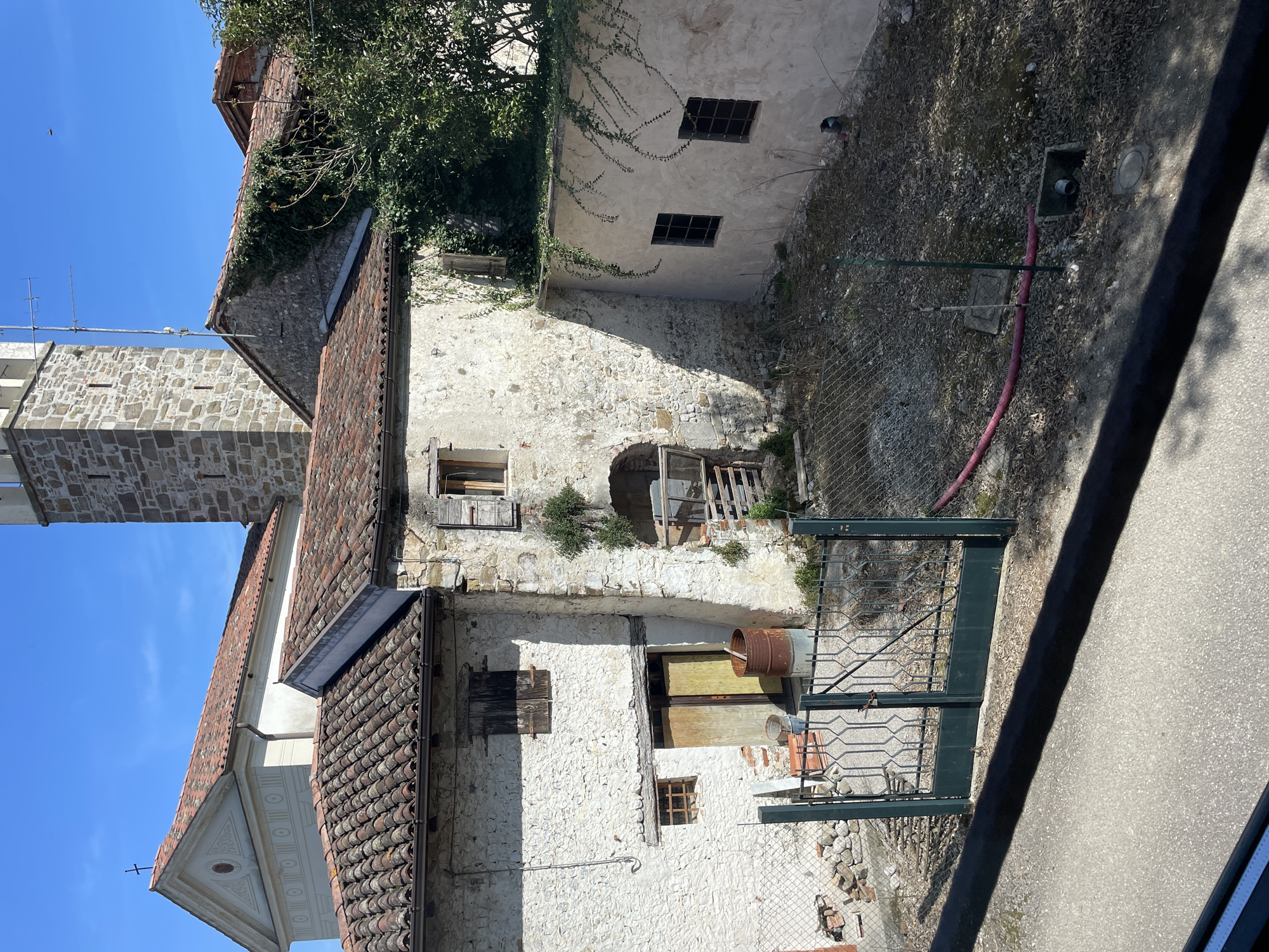 The image shows a weathered, multi-story building with a mixture of stone and plastered walls, located in San Lorenzo, Italy. The building has several windows, some with closed wooden shutters. A rusty metal barrel sits near an open doorway on one of the levels. A green metal gate is visible in the foreground, behind which is a fenced area with some debris. To the left, a tiled roof and a tower are visible, suggesting the building is part of a larger complex or connected to other structures. The sky is clear blue. The overall scene suggests a historical or older part of the city.