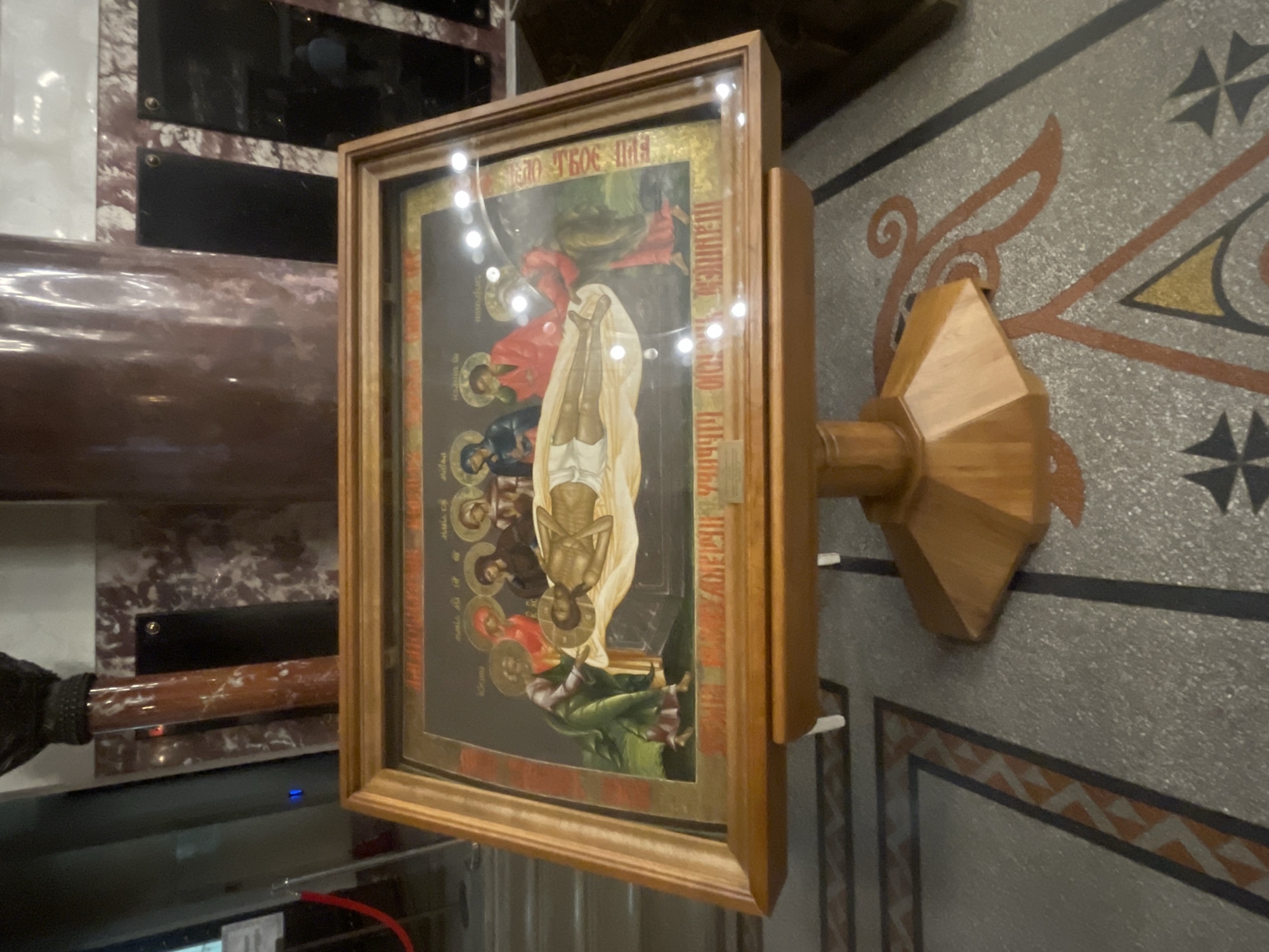This image shows an icon depicting the Entombment of Christ, displayed on a wooden stand in what appears to be an Orthodox church. The icon is framed in wood and placed on a stand with an octagonal base. The scene on the icon features Christ's body lying on a slab, with several figures surrounding him, likely mourners or disciples. The surrounding environment includes columns with a marble-like finish and a patterned floor. A red velvet rope barrier is visible in the background, suggesting a public or accessible area within the church. The lighting in the scene creates reflections on the glass protecting the icon. The location is identified as Kronstadt, Russia.