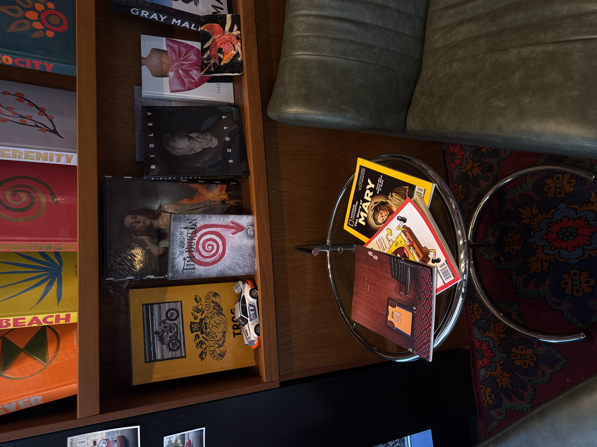 The image captures a scene within a likely commercial or residential interior. A tall, wooden shelving unit displays a collection of oversized coffee table books with titles like "Serenity," "Beach," and "Locity" visible. Further along, are others with names "Ridley Scott" and "The World of Tim Burton." A miniature toy car sits atop one of the books. Next to the shelves is a green leather sofa. A round, multi-tiered glass table is placed between the shelving unit and sofa, holding several magazines with titles such as "Mary" and a book with a picture of a Kobe Bryant jersey. The scene is captured from above, casting shadows that indicate a light source nearby. A patterned rug partially occupies the lower right of the frame. There's nothing to indicate the image was taken in Moscow, Russia.