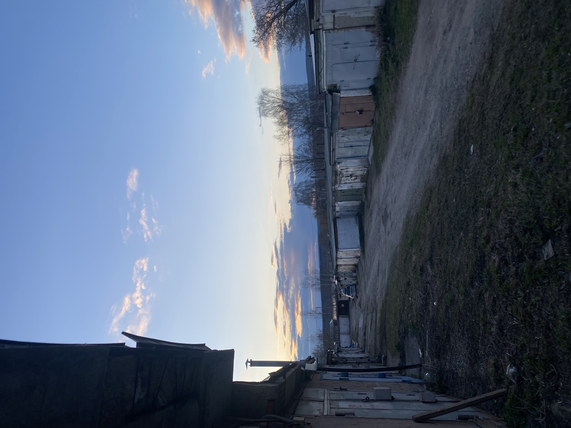 The image depicts an outdoor scene at either sunrise or sunset in Chaikovsky, Russia, showcasing a row of utilitarian garages or storage units. The sky, dominating the upper left, displays a clear blue gradient with scattered clouds. Towards the horizon, these clouds are tinged with warm golden and pink hues, indicative of low sun angle.
Along the right side, a long line of weathered, box-like structures, serving as garages or storage units, stretches into the distance. These structures exhibit varying materials, including corrugated metal and painted wood, showing signs of age and wear. A dirt path runs parallel to this row of garages, separating them from an unkempt strip of grass and weeds on the far right.
In the middle ground, bare trees are silhouetted against the bright sky, suggesting late autumn or early spring. On the left foreground, a darker, less distinct structure with an uneven roofline and a visible chimney or vent pipe is partially visible. Faint outlines of vehicles can be seen parked further down the row of garages. The scene is devoid of human activity, conveying a quiet, somewhat stark atmosphere. No visible text is present.