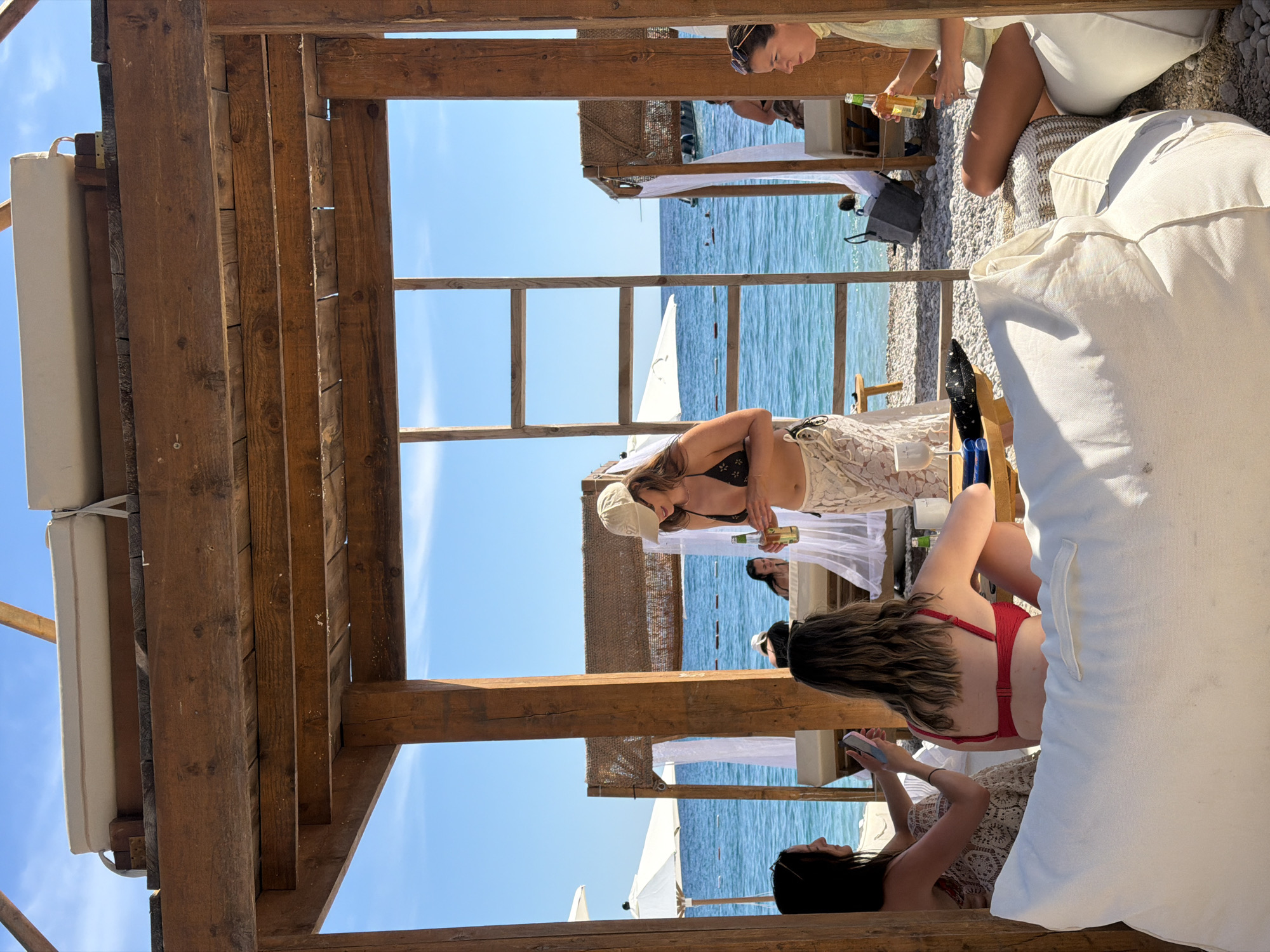 The image shows an outdoor scene at a beachside location, specifically in Sveti Stefan, Montenegro. The foreground is dominated by a wooden structure, likely part of a beach cabana, with white cushions and a lounger. Several women are visible in the scene. One woman, wearing a red bikini top, is lying down and looking at a device, possibly a phone. Another woman, wearing a bikini top and a hat, is standing and holding a bottle. The background features the Adriatic Sea, beach umbrellas, and other similar cabanas. The sky is clear and blue.