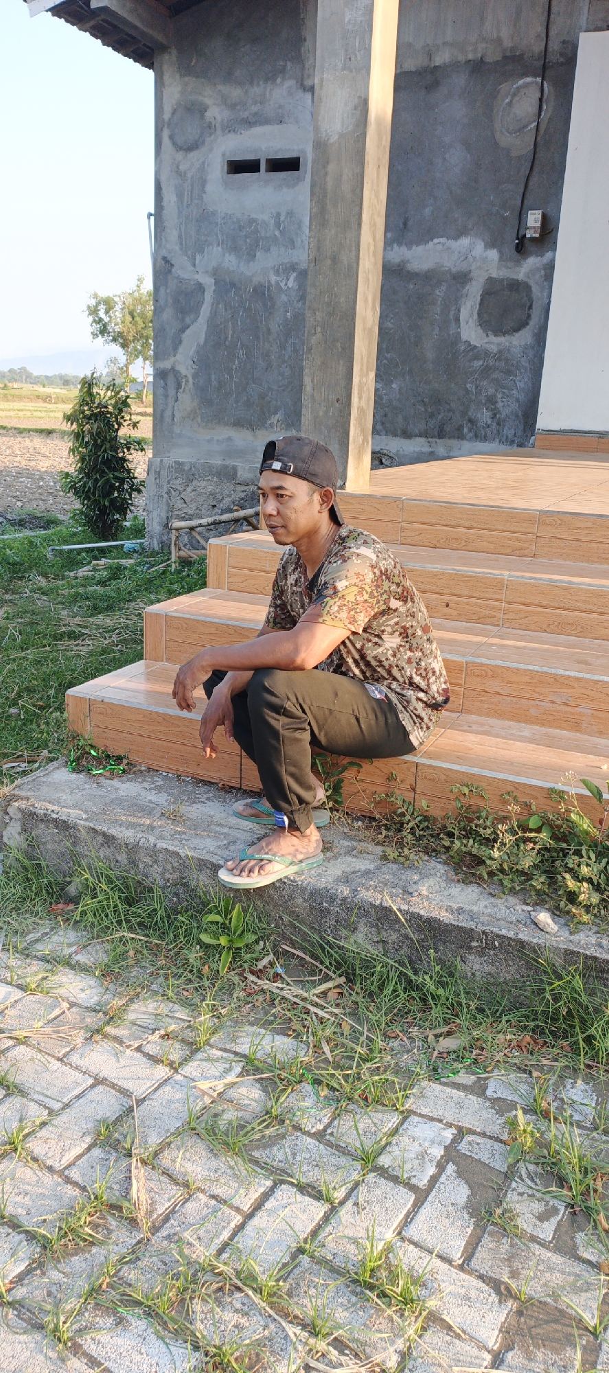 A man is squatting on the steps of a building under construction. He is wearing a camouflage t-shirt, black pants, a black baseball cap, and flip-flops. The building appears to be made of concrete and has a rough, unfinished texture. There are two rectangular openings on the wall of the building, possibly vents. In the background, there is a field with some trees. The ground in front of the steps is paved with interlocking bricks and covered with grass. The lighting suggests it is daytime, possibly late afternoon due to the long shadows. The image is taken from a medium distance, showing the man and his immediate surroundings. The location is identified as Jambearum, Indonesia.