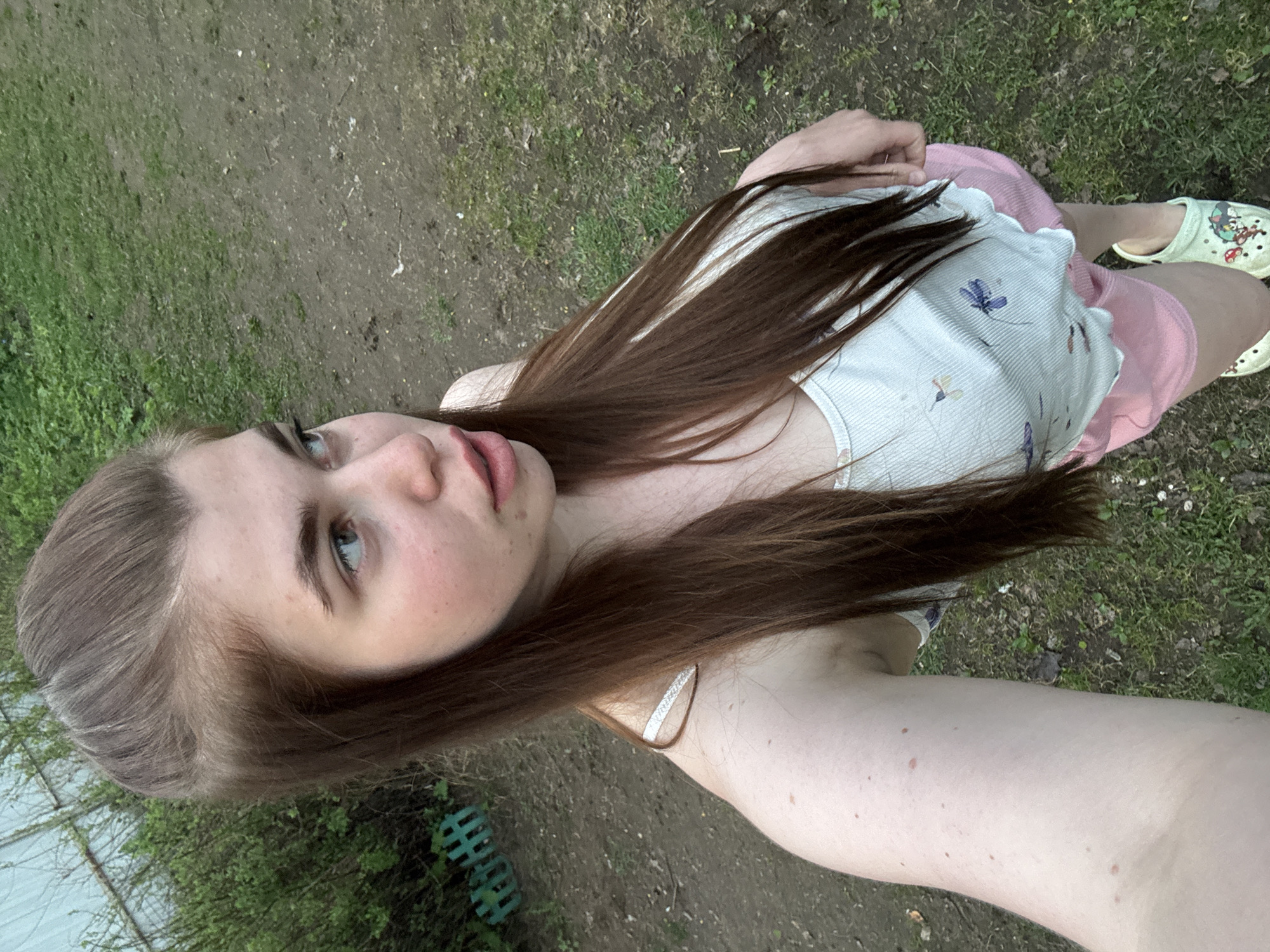 A young woman is outside on a patch of grassy and dirt ground. She's wearing a white tank top with floral print, pink shorts, and light green clogs. Her hair is long and brown, and she is looking upwards and slightly to the left. The background consists of grass, dirt, and some foliage near a weathered structure that could be a fence. The location is not discernable from the image, and therefore cannot be identified as being in the city of Мещёры, Russia.