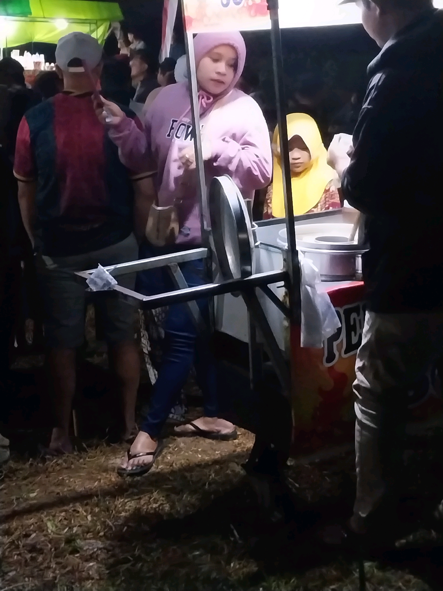 In Nglegok, Indonesia, a group of individuals are gathered around a food cart at night. The cart is adorned with colorful decorations, including the letters "PE" prominently displayed. A young person wearing a pink hooded jacket and blue jeans is seen interacting with the cart, holding a piece of food. Nearby, another person is visible wearing a yellow headscarf. The ground around the cart is covered in dry grass and leaves. The lighting suggests an outdoor evening setting, possibly at a market or fair.