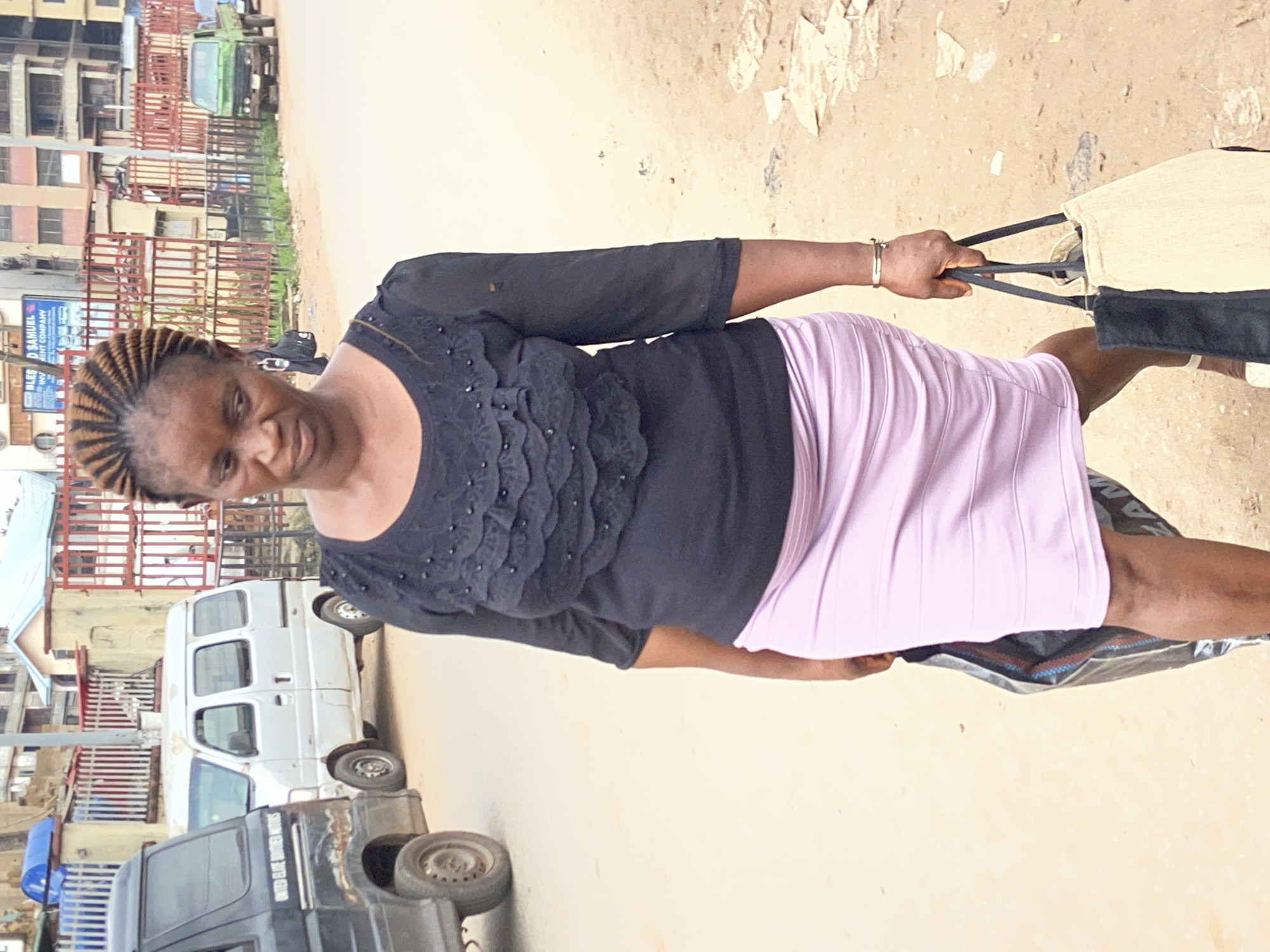 A woman with braided hair walks down a dirt road in Nkpor, Nigeria, carrying two shopping bags. She wears a black, ruffled blouse and a lavender skirt. To her left, a white van and a black truck are parked. In the background, buildings and a green truck can be seen. The woman's gaze is directed downward and slightly to her left. The road is unpaved and dusty, with scattered debris.