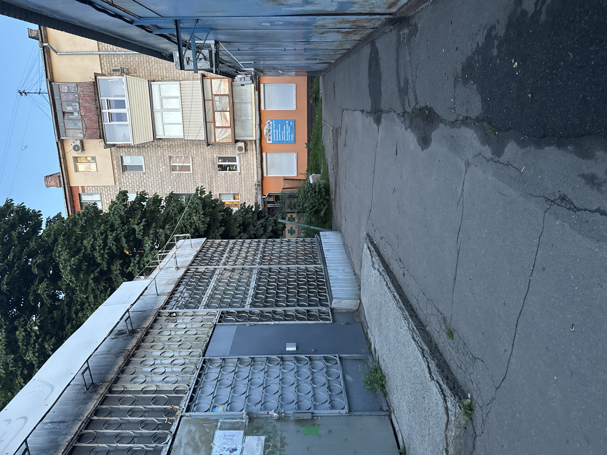 The media file depicts an outdoor scene looking down a narrow paved pathway between two buildings. The pathway is cracked and wet in places, suggesting recent rain or a damp environment.
On the left side of the pathway is a multi-story brick building with several windows and balconies. Some balconies are enclosed with glass, while others have visible air conditioning units. Trees with dense green foliage are growing in front of this building. Above the brick building, a portion of a lighter-colored building with a metal roof is visible.
To the right of the pathway, there is an orange-colored building with two roller shutters. A blue sign with white text and graphics is mounted on this building between the shutters. A glimpse of a metal structure, possibly a fence or awning, runs along the top of the frame on this side.
Further to the right, the pathway is bordered by a textured concrete wall. The overall scene appears to be an urban or suburban setting, possibly an alleyway or service area. The lighting suggests it is daytime, but the sky is not clearly visible. There are no people in the frame, and the scene is static. The metal structures in the foreground on the left have a repeating circular pattern. The text on the blue sign is not fully legible but appears to be in Cyrillic script, possibly listing services.