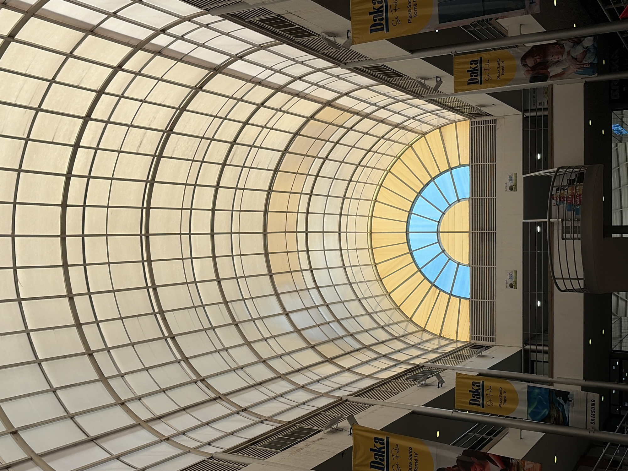 The image, originally rotated ninety degrees counter-clockwise, displays the interior of a large building, likely a shopping mall, in Ciudad Guayana, Venezuela. The central feature is a massive, curved skylight structured with a grid of translucent panels that cast subtle shadows. A prominent semi-circular section of the skylight is distinctly colored in blue and yellow, allowing a direct view of a clear sky and indicating daytime.
Vertically hung banners are visible along the building's multi-level interior. Multiple banners prominently feature the brand "Daka" and the Spanish phrase "Se Feliz" (Be Happy), along with the location "Plaza Santo Tomé IV." Other visible text includes "REGISTRO MAR" on a wall, a "100%" sign, and a partially visible "SAMSUNG" logo on one of the banners. A sign on a lower level also shows "Diseño Graf," likely an abbreviation for "Diseño Gráfico" (Graphic Design). The scene showcases the building's architecture, including railings, ventilation grilles, and embedded lighting fixtures. No people are visible, highlighting the spacious and architectural nature of the setting.