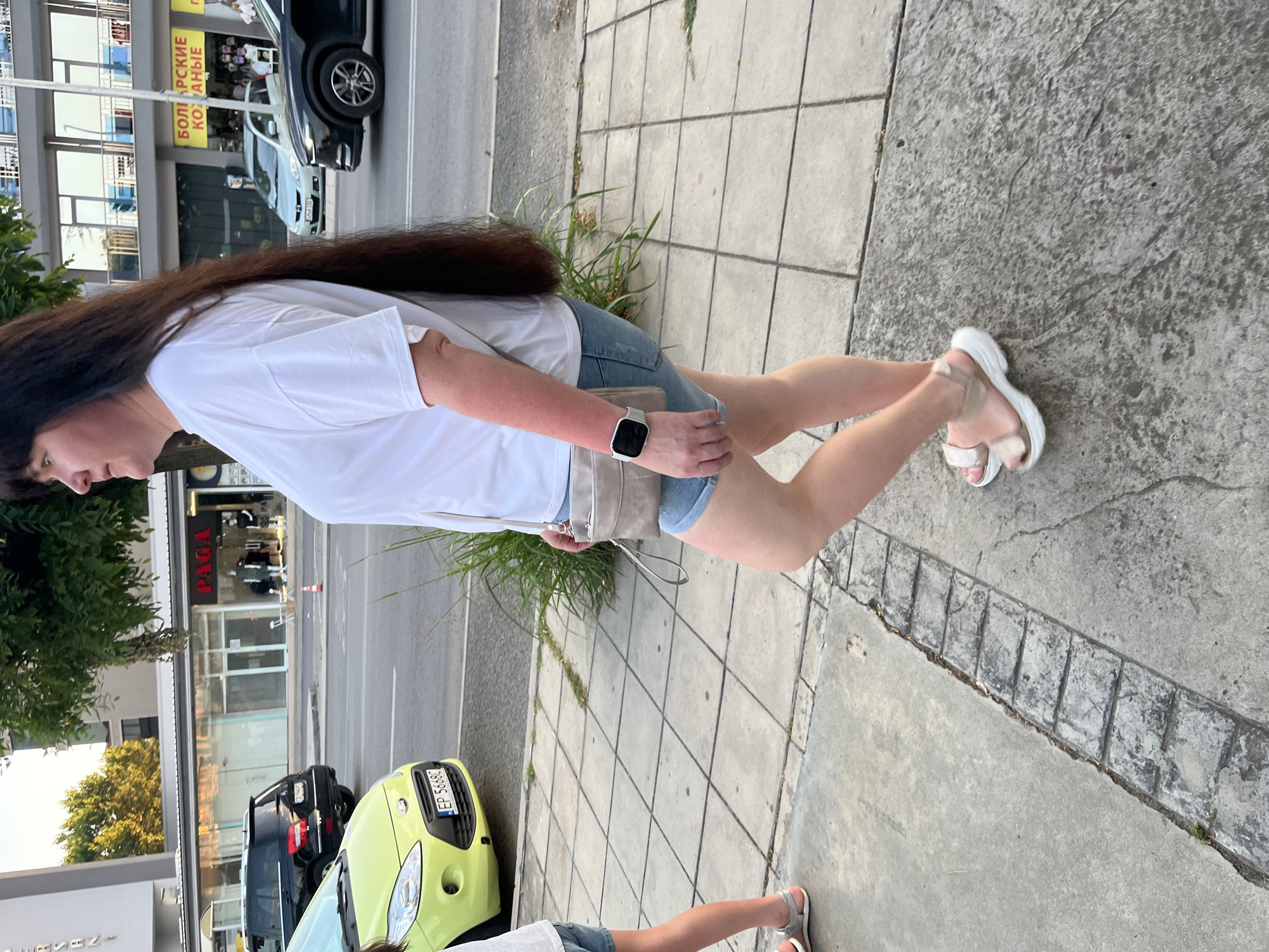 In Nesebar, Bulgaria, a woman with long dark hair is walking on a sidewalk. She is wearing a white t-shirt, denim shorts, and sandals. A light-colored crossbody bag is slung over her shoulder, and she has a smartwatch on her left wrist. The sidewalk is paved with square concrete tiles and bordered by a strip of smaller rectangular stones. Behind the woman, a street runs parallel to the sidewalk, with several cars visible. Businesses line the street, with storefronts displaying signs, including one that reads "PAGA". Greenery and trees are present in the background. The lighting suggests it is daytime.