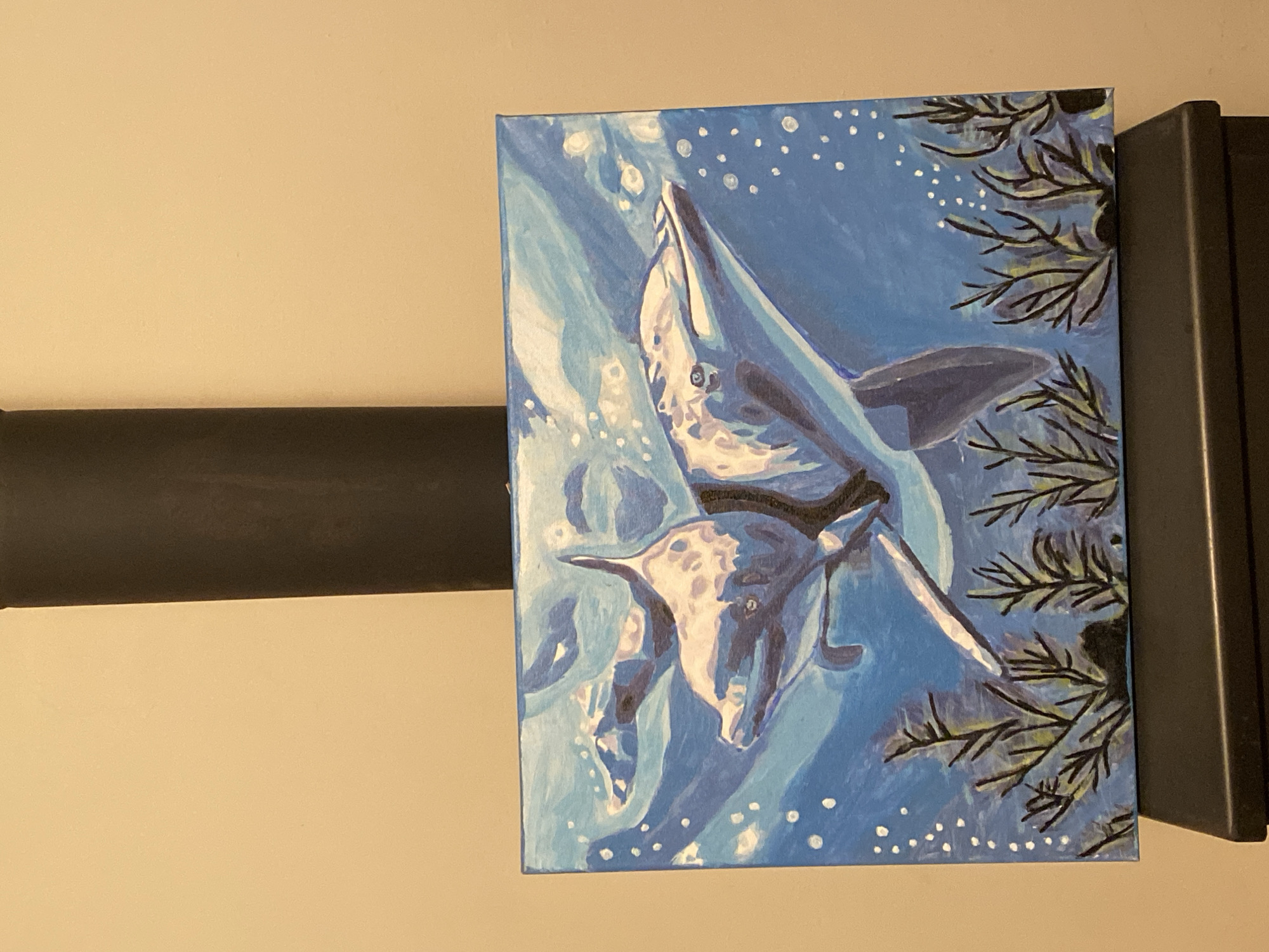 A painting depicts a marine scene in shades of blue. Two manta rays are depicted, one larger than the other, swimming upwards. White dots, likely representing bubbles, are scattered throughout the blue background. Dark green and black branches with small leaves are painted on the right side of the canvas. The painting is positioned on a flat surface with a dark brown object adjacent to the painting on the left side, and a black rectangular object behind it on the right. The background wall is a light beige color. There is no information to suggest that this image was taken in Goritsa, Bulgaria.
