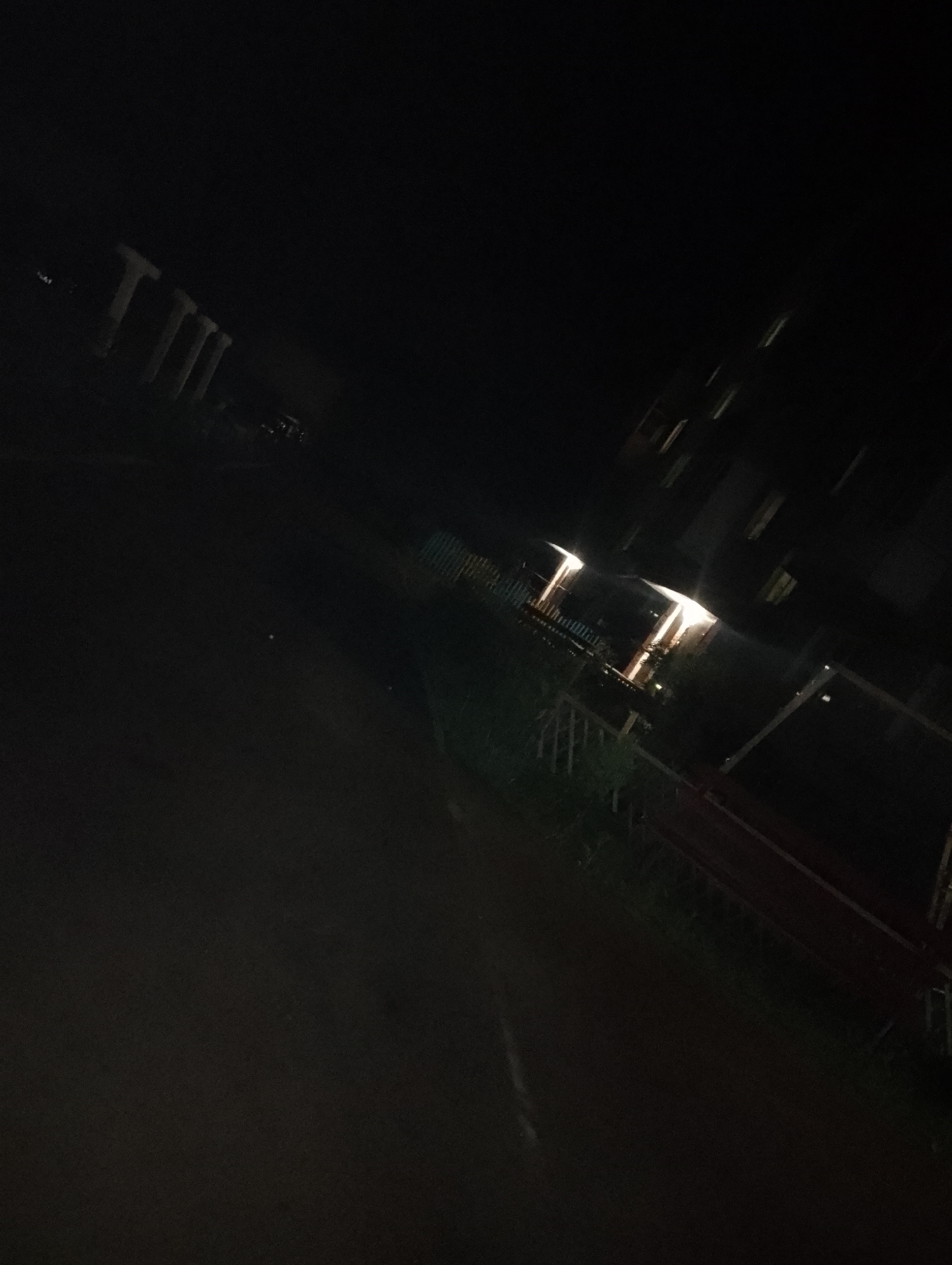 The image depicts an outdoor night scene in an unknown city. A dark, paved road occupies much of the foreground, leading towards a structure lit by warm-toned light. This structure is bordered by a fence and vegetation, suggesting it could be a building or garden. The background features indistinct dark shapes that could be other buildings or trees. The overall impression is of a dimly lit, somewhat deserted urban area.