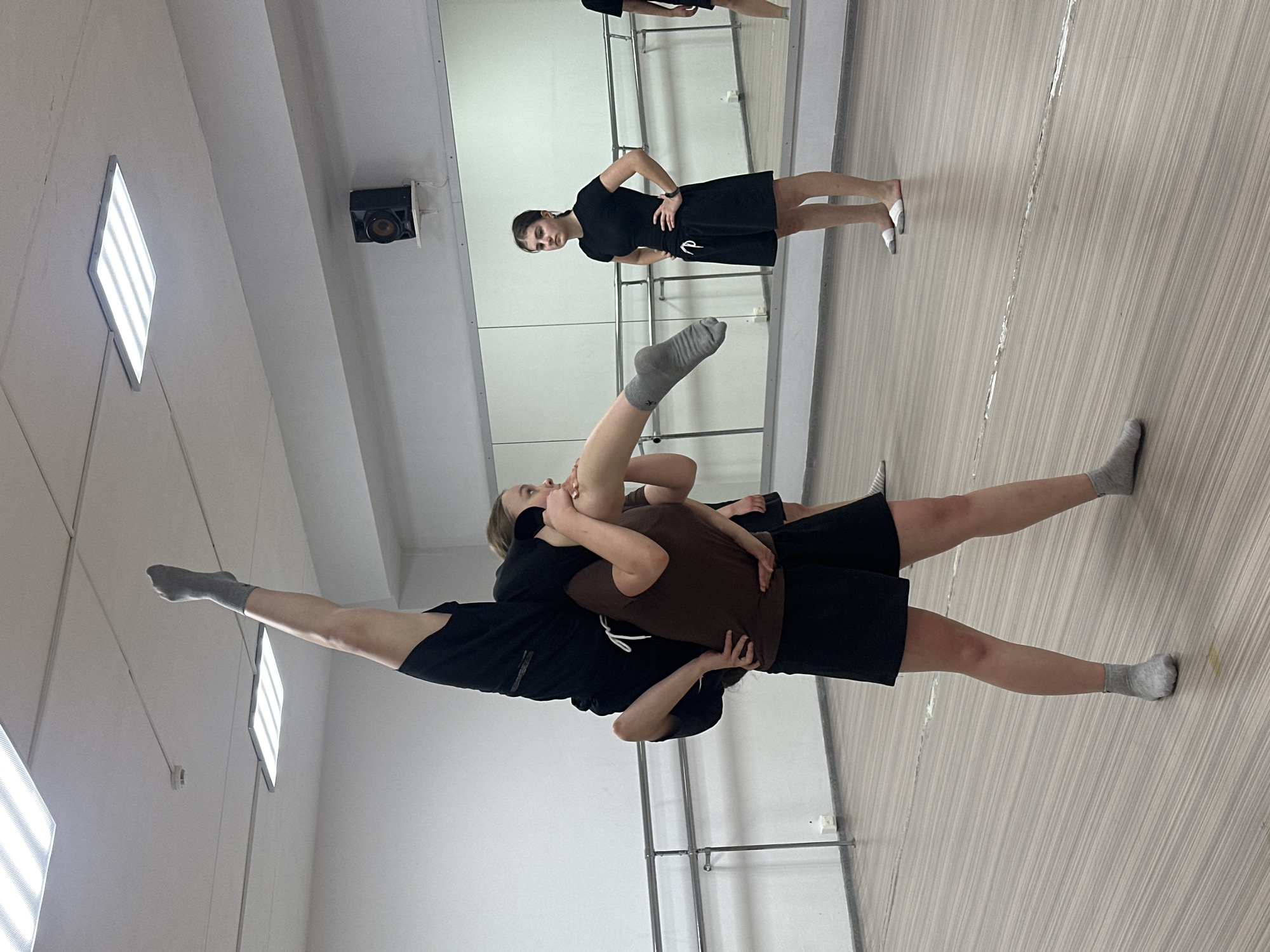The image shows a dance studio. Two women are in the foreground: one woman is holding the other up while the woman being held extends her legs to either side. Another woman is in the background, standing with her hands on her hips, facing the mirror. The studio has a wood-patterned floor and a wall of mirrors. Light fixtures are on the ceiling. The location may be a dance studio in Votkinsk, Russia.