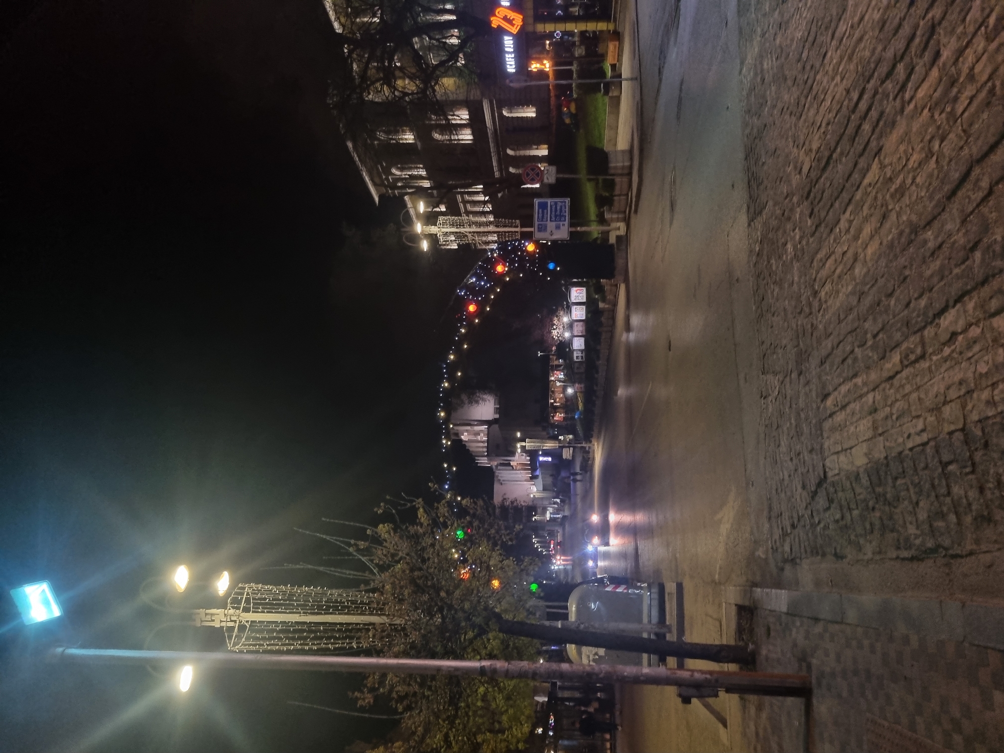 The image depicts a street scene at night in Veliko Tarnovo, Bulgaria. The street is illuminated by streetlights and decorative lights strung across the road. There are buildings on either side of the street, including a building with the sign "Cafe Joy 23" on the upper left side of the scene. A string of colored lights arches over the street, creating a festive atmosphere. Trees with decorative lights are positioned along the roadside. The street surface appears to be a combination of pavement and cobblestones. Cars are visible on the street.