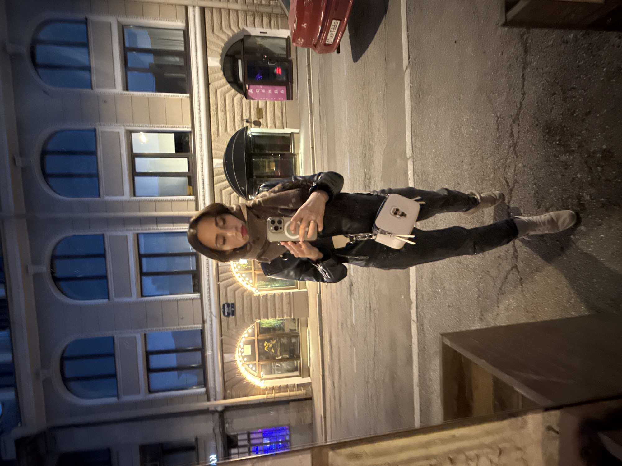 A full-body mirror selfie taken at night on a city street in Saint Petersburg, Russia, shows a young woman with dark hair, wearing a black leather jacket, dark pants, a grey scarf, and light-colored boots. She holds a smartphone in her right hand, capturing her reflection. A small, light-colored purse with a chain strap is worn across her body.
The background reveals a multi-story building with arched windows, some illuminated with festive lights. Across the street, a red car is parked, and further down the street, another building entrance with a glowing sign is visible. The street is paved, with visible white lines and some cracks in the asphalt. The overall atmosphere is urban and dimly lit, characteristic of a nighttime city environment.