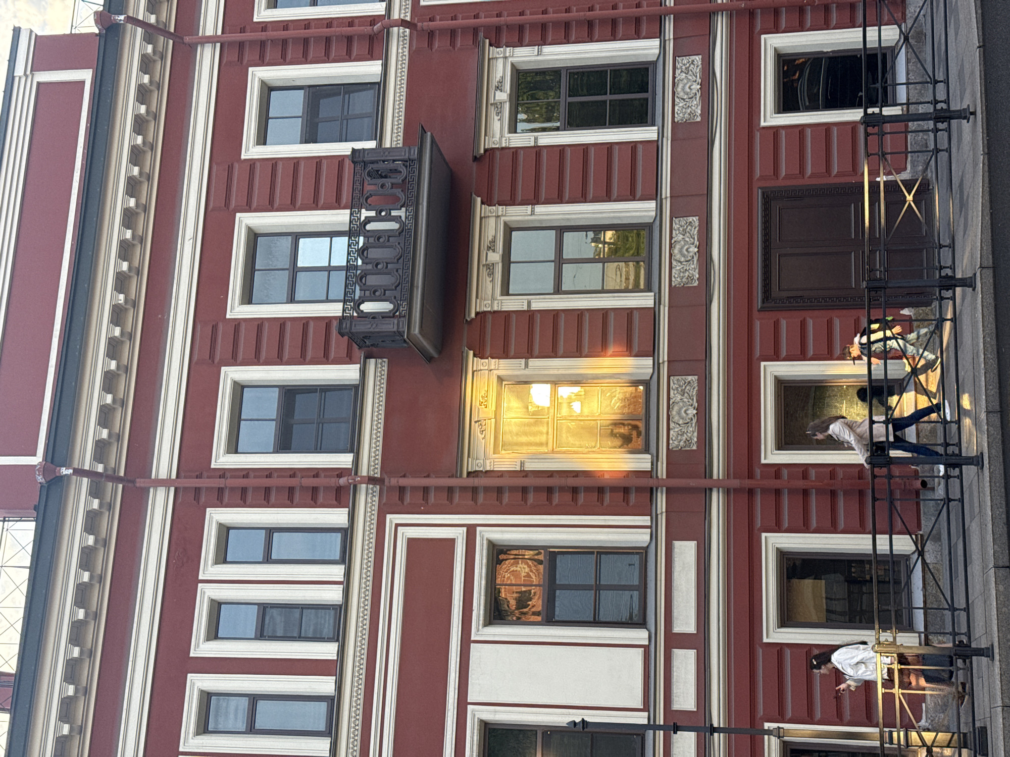 The image shows a section of a building's facade in Saint Petersburg, Russia. The building is painted in a reddish-brown color with white architectural details. Windows of varying sizes are visible, some with dark glass and one reflecting sunlight. A wrought iron balcony projects from the facade. Two people are on a fire escape, one looking down and the other looking at a phone. A drainpipe runs along the length of the building.