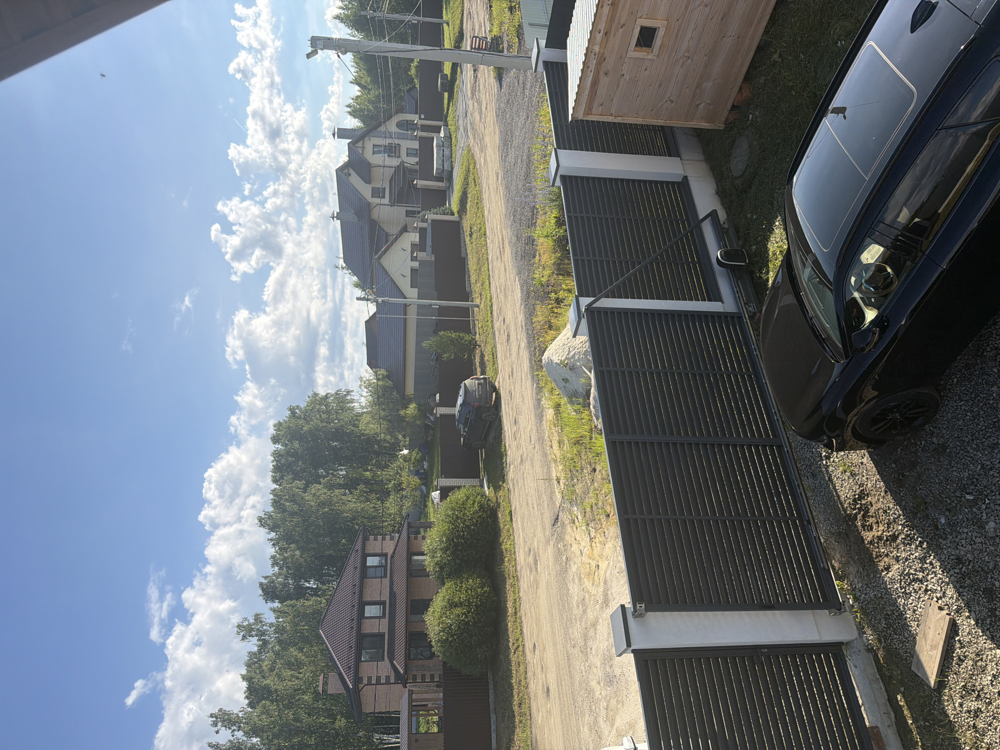 The image depicts a suburban street scene in Malinovka, Russia, bathed in bright sunlight under a partly cloudy sky. In the foreground, a black car with a sunroof is parked on a gravel surface. Adjacent to the car is a modern black metal fence with white-topped pillars, partially obscuring a wooden outbuilding. Beyond the fence, a dirt road winds through the landscape, leading towards a row of houses. The houses are of varying architectural styles, with dark roofs and light-colored walls, surrounded by fences and greenery. A dark-colored SUV is visible parked in front of one of the houses. A line of mature trees forms a backdrop to the scene, suggesting a rural or semi-rural setting. The overall impression is one of a quiet, residential area on a clear day.