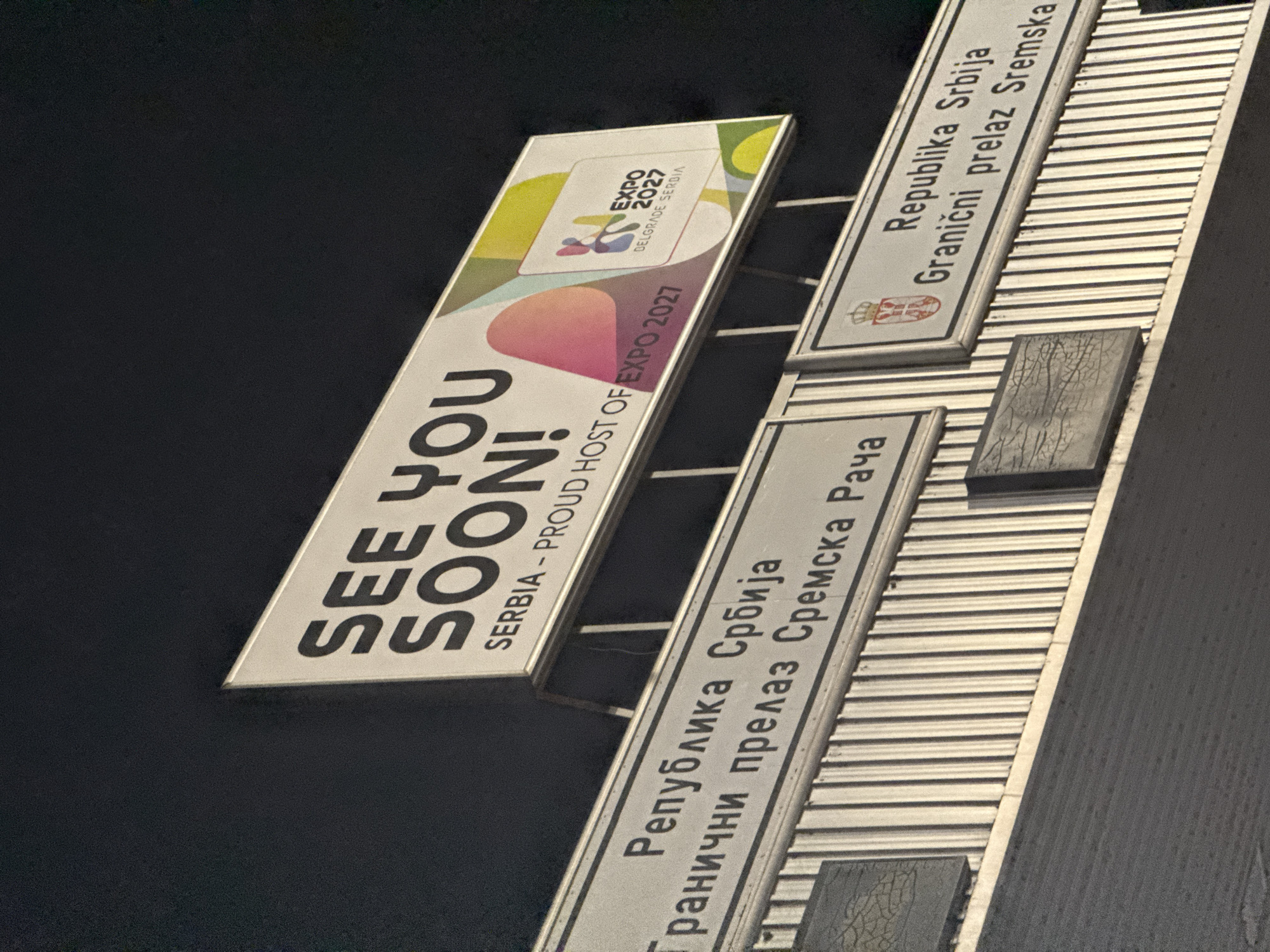The image captures a set of illuminated signs against a dark background, likely at night, indicating a border crossing into Serbia. The primary subject is a large, vertical promotional sign for EXPO 2027. This sign features the text "SEE YOU SOON! SERBIA - PROUD HOST OF EXPO 2027" in bold, white capital letters, accompanied by the "EXPO 2027 BELGRADE SERBIA" logo. The logo itself has an abstract, colorful design with elements in yellow, green, pink, and white.
To the right and below this promotional sign are two horizontal signs, characteristic of a border crossing. The upper border sign clearly reads "Republika Srbija / Granični prelaz Sremska Rača" in Cyrillic script, with the coat of arms of Serbia visible on the left. A lower, partially visible sign repeats "Republika Srbija / Granični prelaz Sremska" in Latin script, also with the Serbian coat of arms.
The setting is the Sremska Rača border crossing in Serbia, indicated by the text on the border signs. No people are visible. The dark background suggests the image was taken at night, with artificial light illuminating the signs. The overall impression is one of arrival and welcome into Serbia, specifically promoting the upcoming EXPO 2027 in Belgrade.