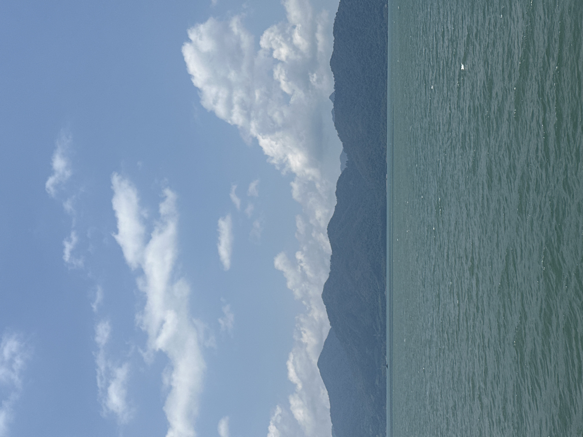 The image shows a coastal landscape in Laem Ngop District, Thailand. The scene is divided into three main sections: sky, land (mountains), and sea. The sky is bright blue with scattered white clouds. The land is a range of dark green mountains. The sea is a calm, blue-green color with a slightly textured surface due to small waves. The photograph is taken on a sunny day.