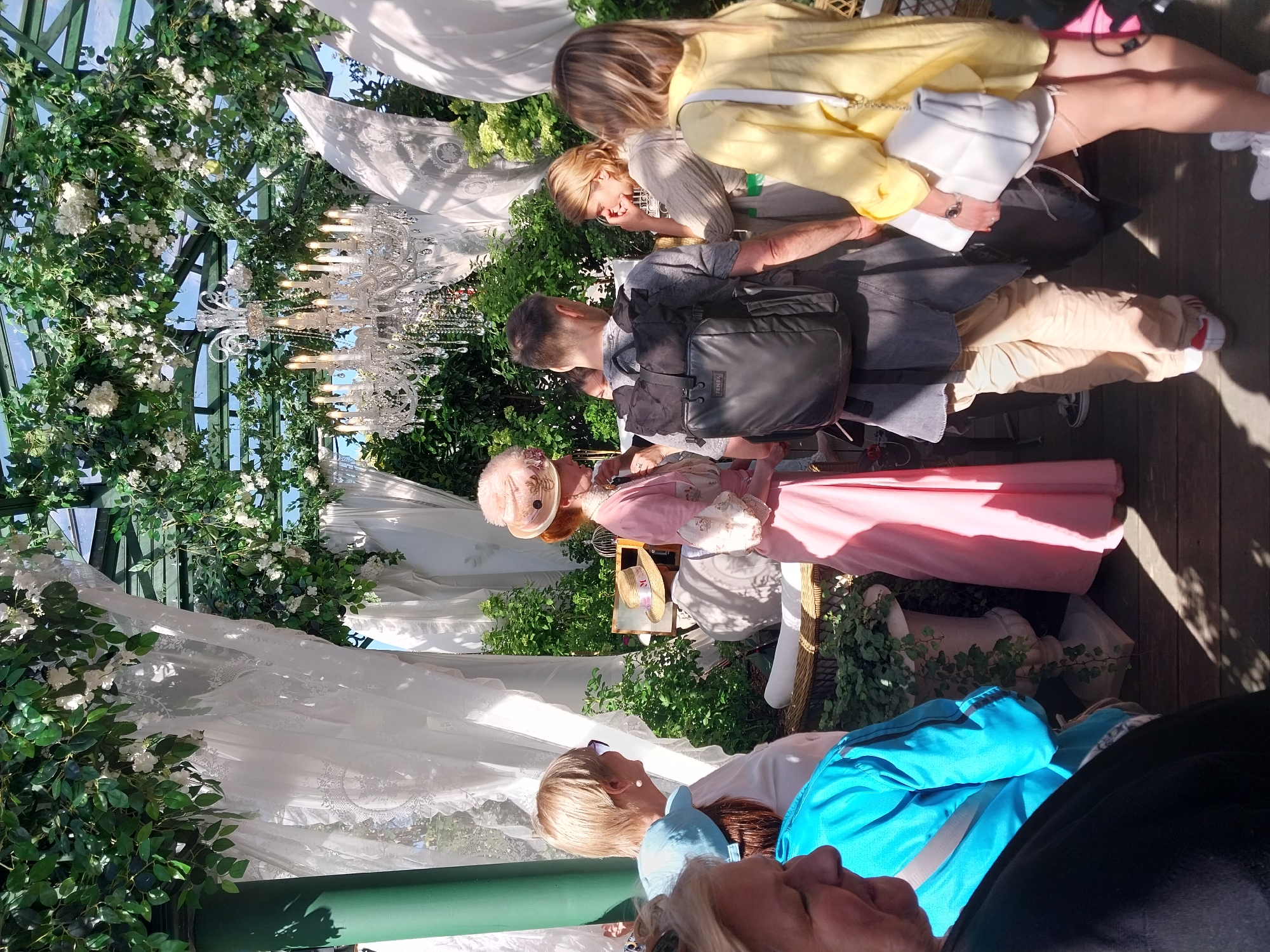 In Moscow, Russia, a group of people are gathered outdoors, possibly at a garden party or an outdoor event. The scene is lush and decorative, with abundant greenery and white floral arrangements. A large, ornate crystal chandelier hangs prominently, suggesting a festive or elegant atmosphere. Several individuals are visible, some in contemporary clothing and others dressed in what appears to be historical or costume attire. One person is wearing a pink, ruffled dress and a feathered hat, while another is in a yellow jacket and white handbag. The presence of a large backpack on one of the individuals hints at a casual element within the otherwise elaborate setting. The lighting suggests it is daytime, with sunlight casting shadows on the wooden floor. The mood appears relaxed and social, with people interacting and observing.