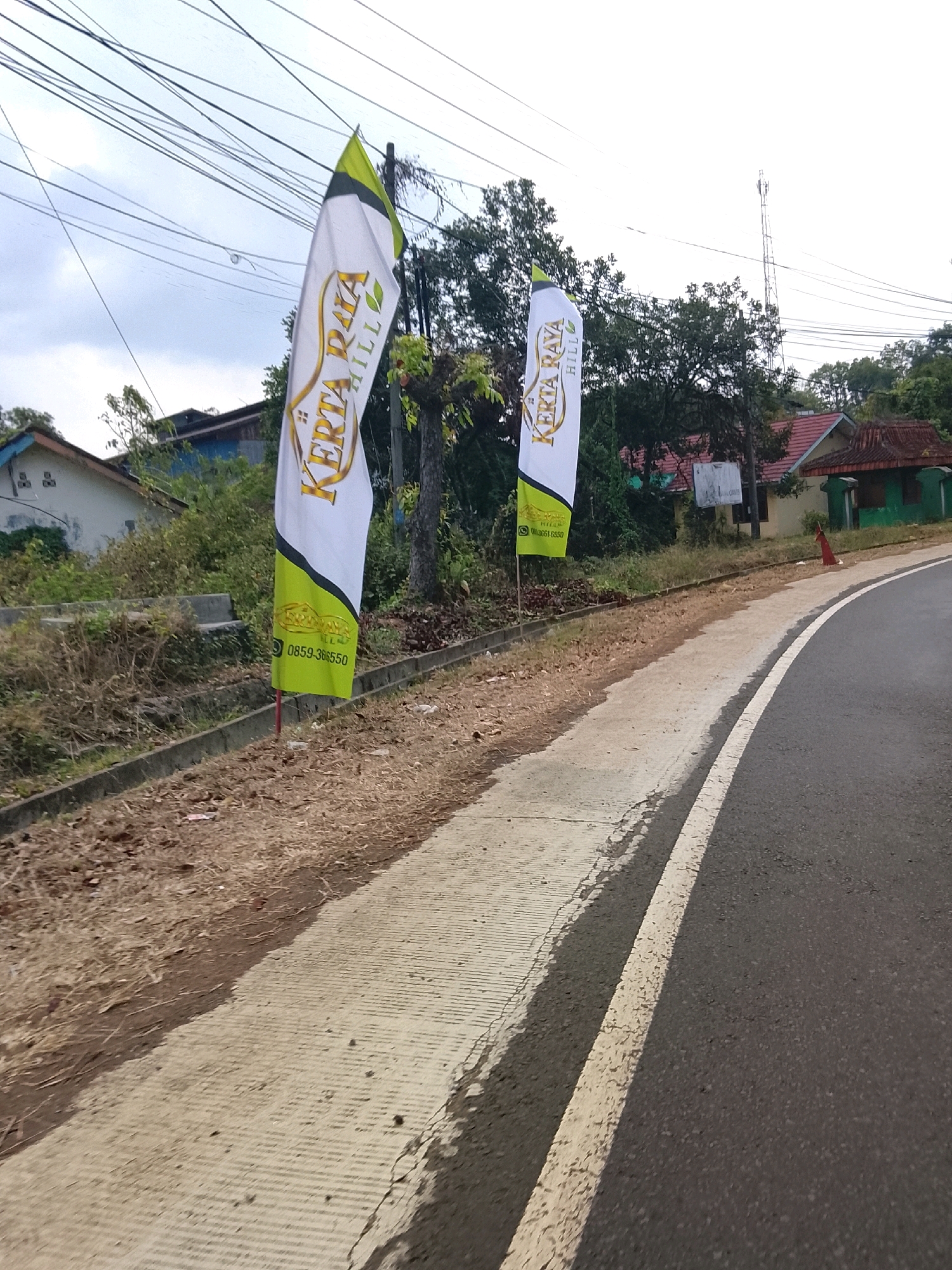 Two tall, vertical banners are displayed on the left side of a paved road. The banners are white with green and yellow accents and feature the text "KERTA RAYA HILL". One banner has the phone number "0859-366550" visible on it. The road is lined with a concrete sidewalk and a dirt embankment covered with dry grass and some vegetation. In the background, there are several houses, trees, and electrical wires. A cell tower is visible in the distance. The sky is overcast. This scene is located in Banjar, Indonesia, specifically along the road leading to the Kerta Raya Hill housing complex.