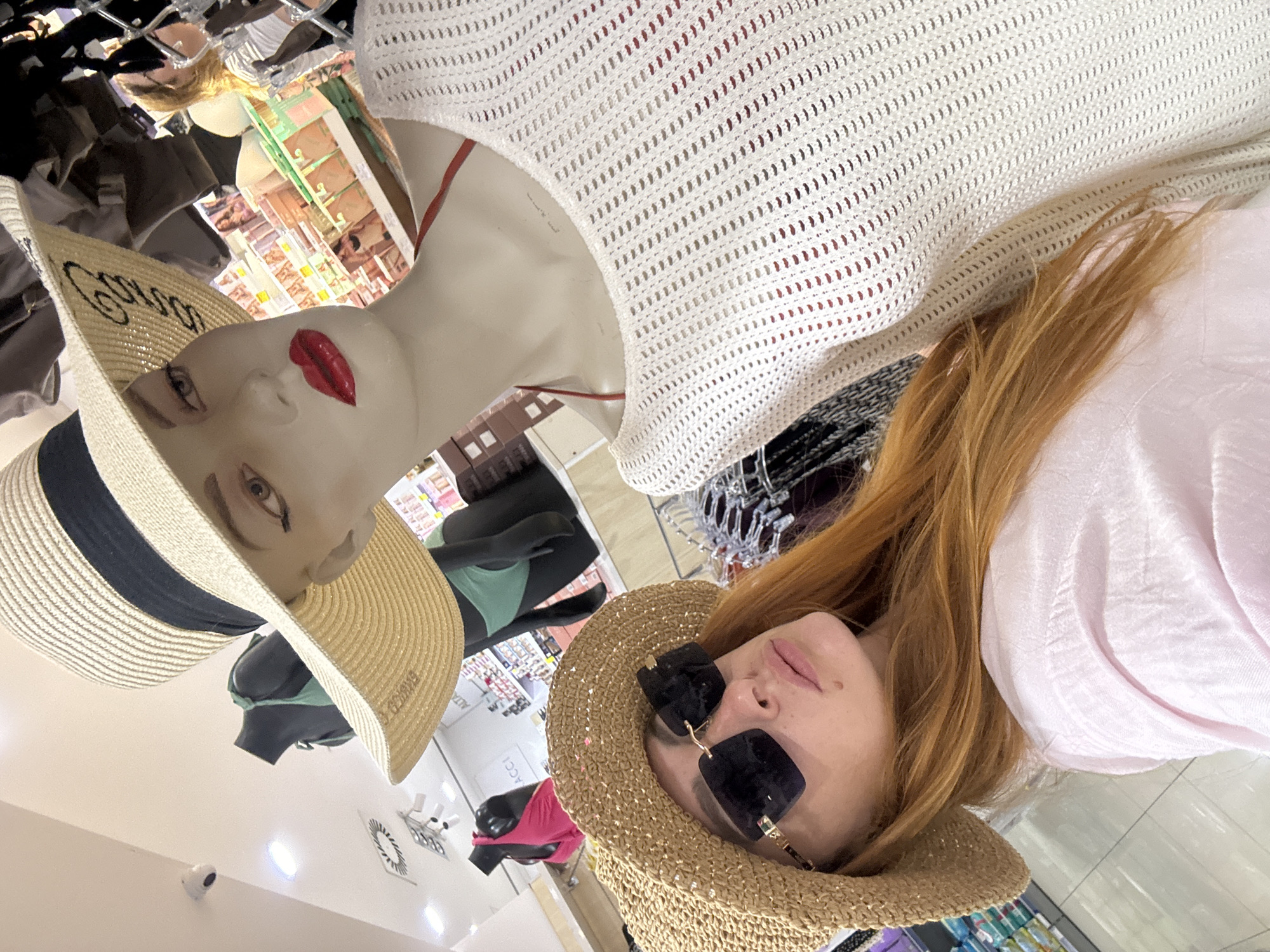 A woman with long, red hair and sunglasses is taking a selfie in a store in Budva, Montenegro. She is wearing a straw hat and a light pink shirt. In the background, a mannequin is wearing a large straw hat and a white knitted top. The store appears to be a clothing or souvenir shop, with racks of clothing and shelves stocked with merchandise visible.