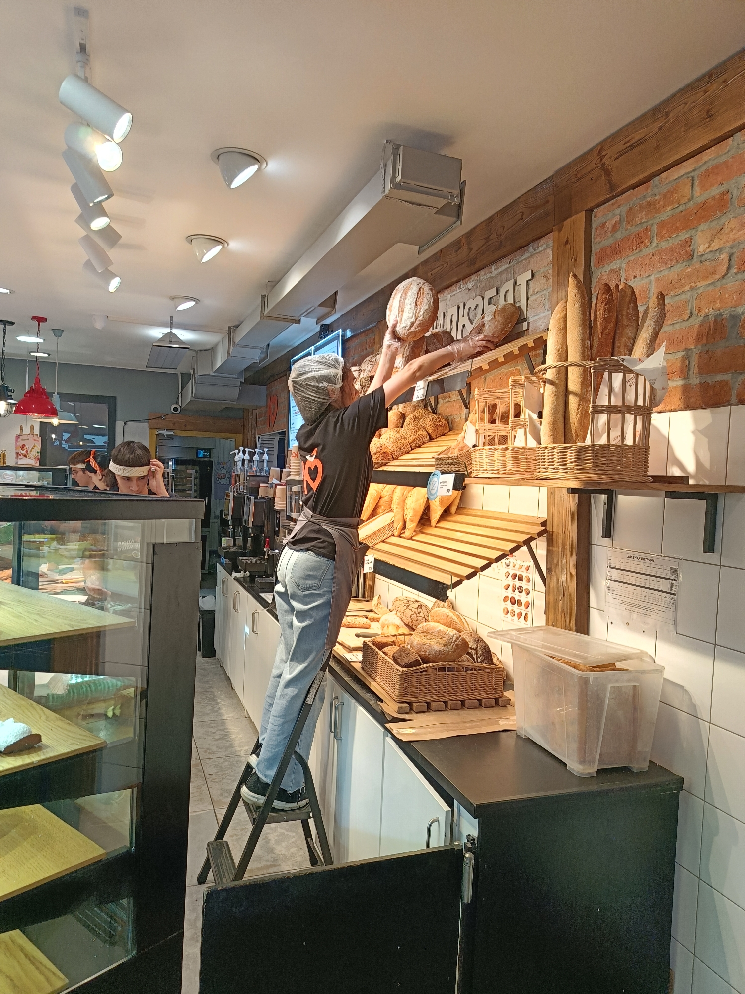 A person, likely a baker or shop employee, is standing on a step stool reaching up to adjust loaves of bread on a high shelf in a bakery. They are wearing a dark t-shirt with an orange heart logo, a grey apron, and light blue jeans. Their hair is covered with a transparent hairnet. Several other people are visible in the background, one behind a glass display case and others in what appears to be a kitchen area.
The scene is set inside a bakery with exposed brick walls and wooden shelving units displaying a variety of bread products, including baguettes and round loaves. There are also wicker baskets and a clear plastic container filled with bread. The lighting is a mix of track lighting on the ceiling and spotlights directed at the merchandise. To the left, there's a refrigerated display case filled with baked goods.
The time of day appears to be during business hours, given the presence of customers or other staff. There are no clear indications of weather. The overall atmosphere suggests a busy but organized retail environment.
Visible text includes: "МУЖСКАЯ ВЫПЕЧКА" (Men's Baking) on a sign above the bread display, and what appears to be a price list or product information sheet on the wall to the right, with Russian text like "ХЛЕБНАЯ ВИЗИТКА" (Bread Business Card).