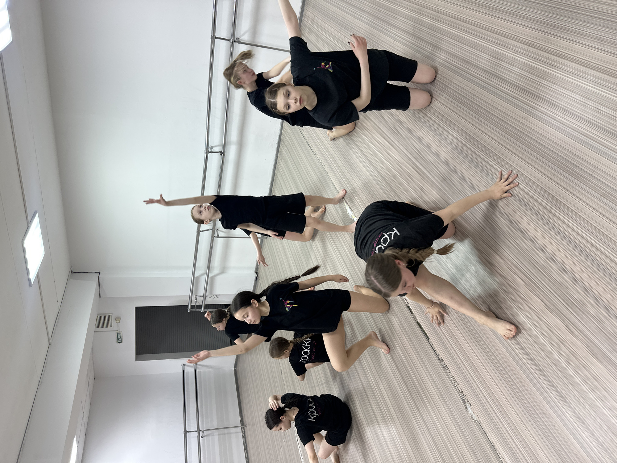 The image captures a dynamic scene of several young girls or teenagers engaged in a dance practice session within a brightly lit studio. The photograph is taken from a high, diagonal perspective, looking down at the performers. There are approximately seven to eight individuals, all dressed uniformly in black t-shirts and black shorts, with most having their hair tied back in braids or ponytails, and performing barefoot.
The girls are in various poses indicative of contemporary or modern dance, often involving floorwork. Some are on their knees with arms extended, others are in low, crouched stances, and a pair in the upper right appears to be in a close, supportive pose, possibly a lift or partner work. Their faces show expressions of concentration and focus as they execute their movements.
The setting is a clean, well-maintained dance studio featuring light-colored, striped wooden floorboards and plain white walls. Along the walls are chrome ballet barres or practice rails. In the background, rectangular ceiling lights illuminate the space, and a ventilation grate is visible. A prominent green sign with the word "ВЫХОД" (meaning "Exit" in Russian) is affixed to a distant wall, confirming the Russian context of the location (Votkinsk). On some of the black t-shirts, the text "КРОКУС" (Crocus) is visible, with smaller text underneath, indicating "dance studio," suggesting it is the name of their dance group or academy. The overall scene conveys a sense of dedicated and artistic training.