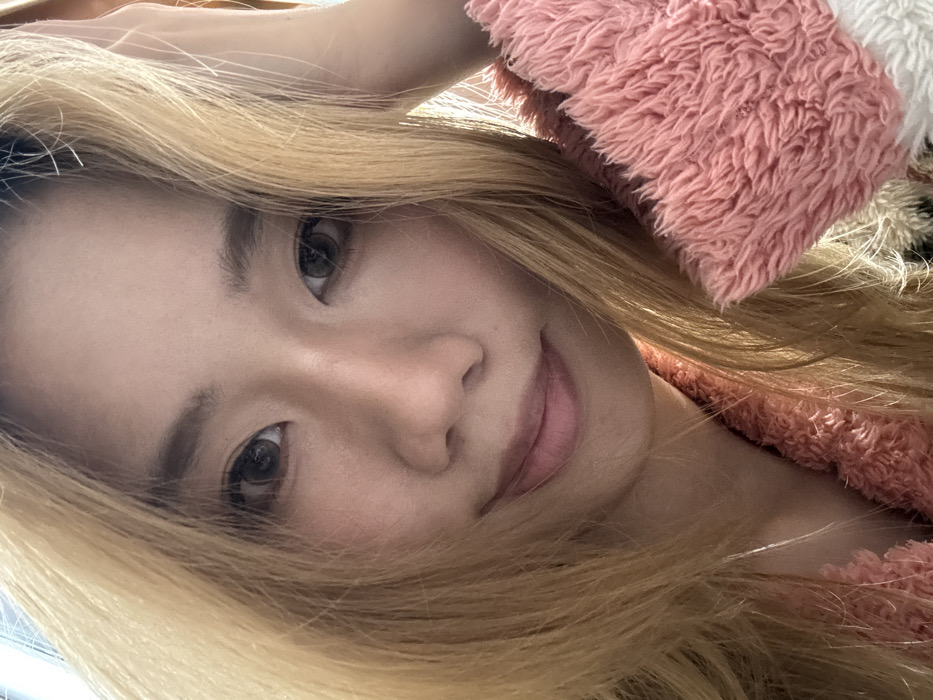 A single East Asian female subject is depicted in a close-up, headshot-style portrait, located in Shapingba District, China. The subject has long, straight, dyed blonde hair, brown eyes, and light skin. She is looking directly at the camera with a slight, closed-lip smile. Her right hand is partially visible, positioned near the right side of her head, and she is wearing a textured, fuzzy pink garment, possibly loungewear. The lighting is soft and frontal, illuminating her face and hair. The background is indistinct and light-colored.