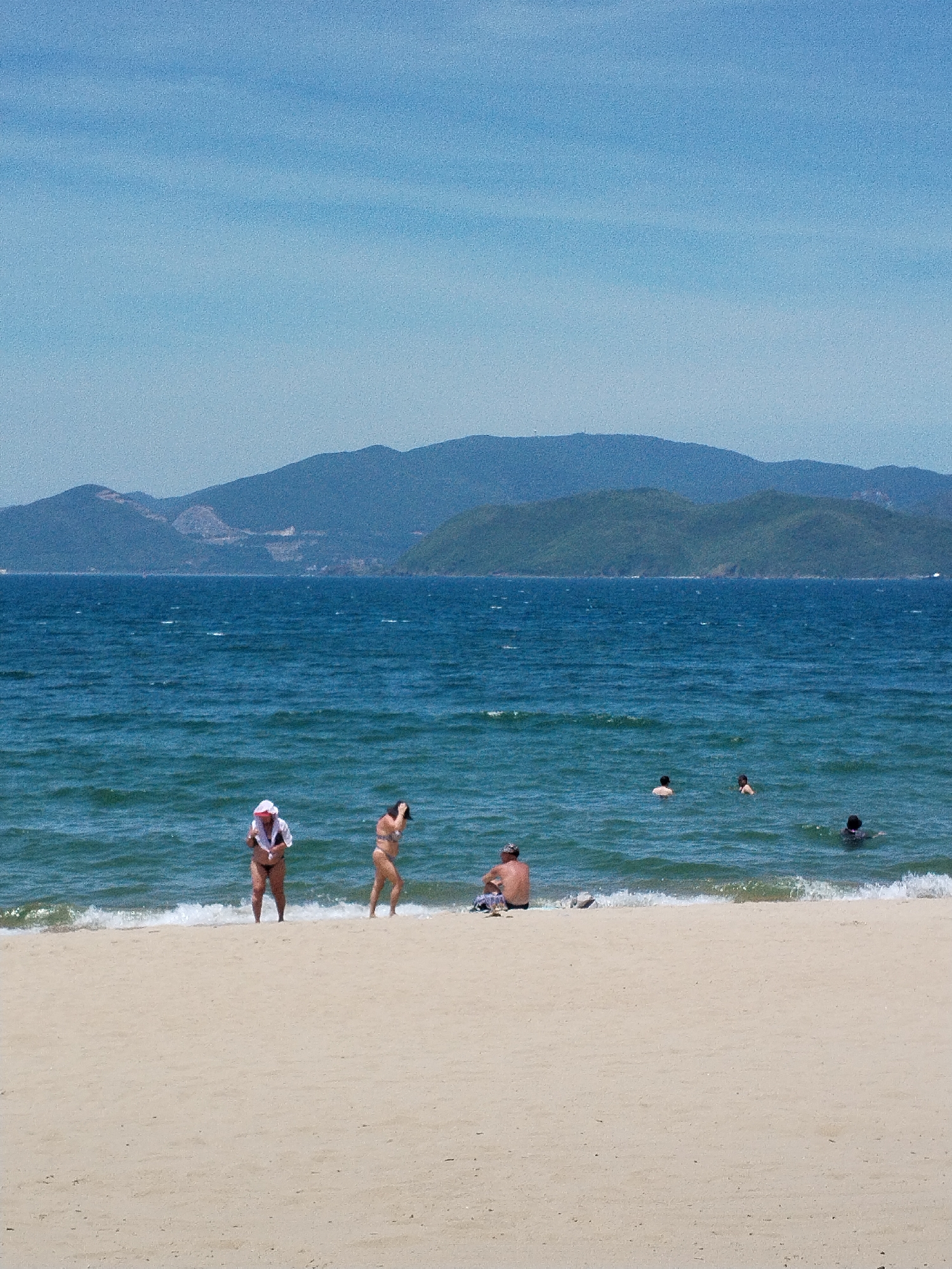 The image depicts a beach scene in Nha Trang Ward, Vietnam. The sand is light beige, and gentle waves are breaking on the shore. In the foreground, on the sand, a person is sitting. Further back, near the water's edge, two women are standing. One is wearing a white headscarf and a bikini, while the other is in a bikini. Several individuals are swimming or wading in the bright blue ocean. In the background, a series of lush green mountains with some lighter patches, possibly indicating structures or cleared areas, rise against a clear, pale blue sky. The overall atmosphere is tranquil and suggests a sunny day at the beach.